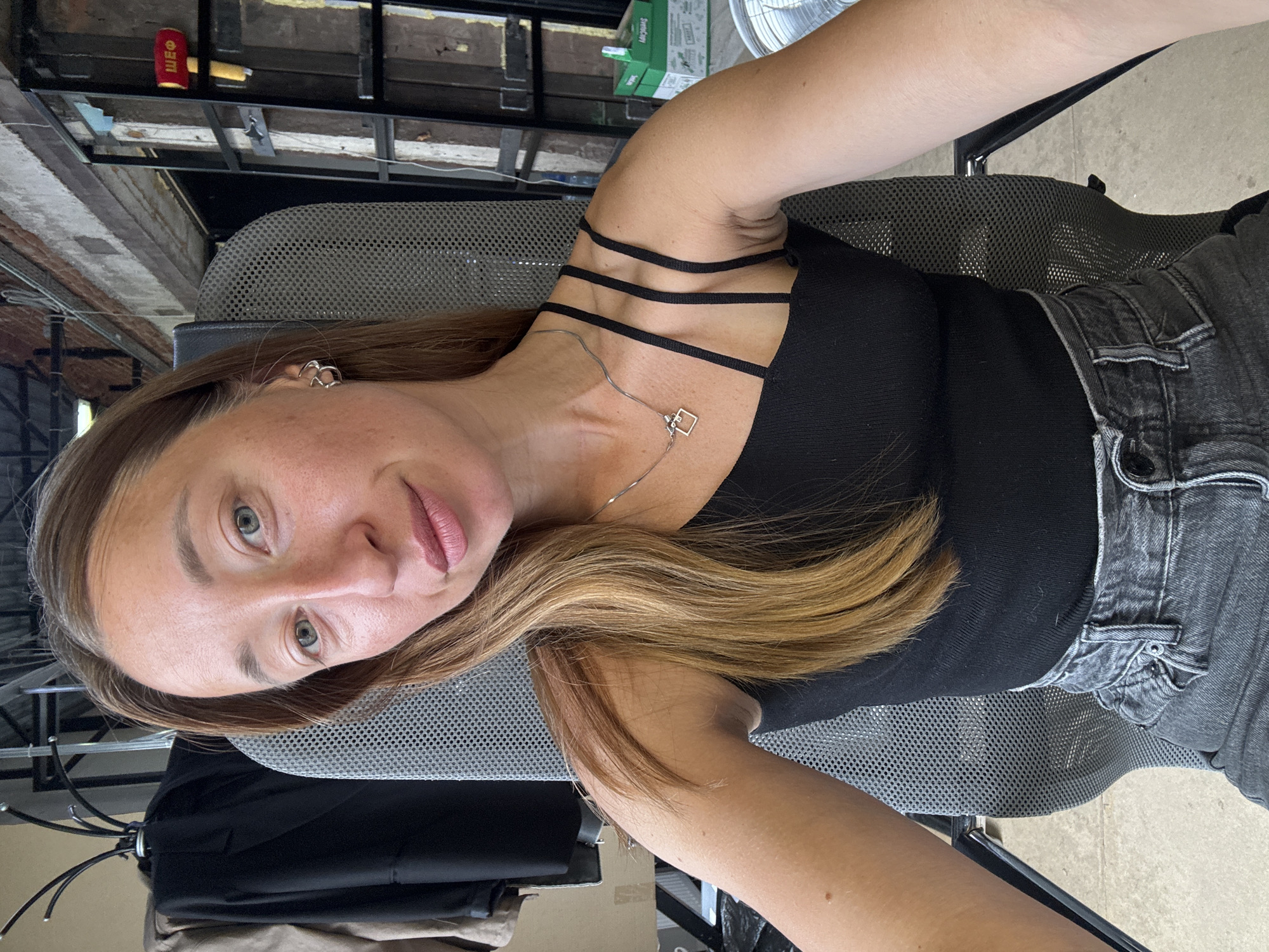 A medium shot depicts a woman sitting in a gray mesh office chair, indoors. She wears a black tank top with multiple thin straps and gray jeans. A silver necklace with a square pendant is visible. Her hair is light brown with blonde highlights. Her makeup is minimal. The background includes a brick wall and industrial-style metal beams. A red can labeled "ШЕФ" can be seen. The image was taken in the city of Chaikovsky, Russia.