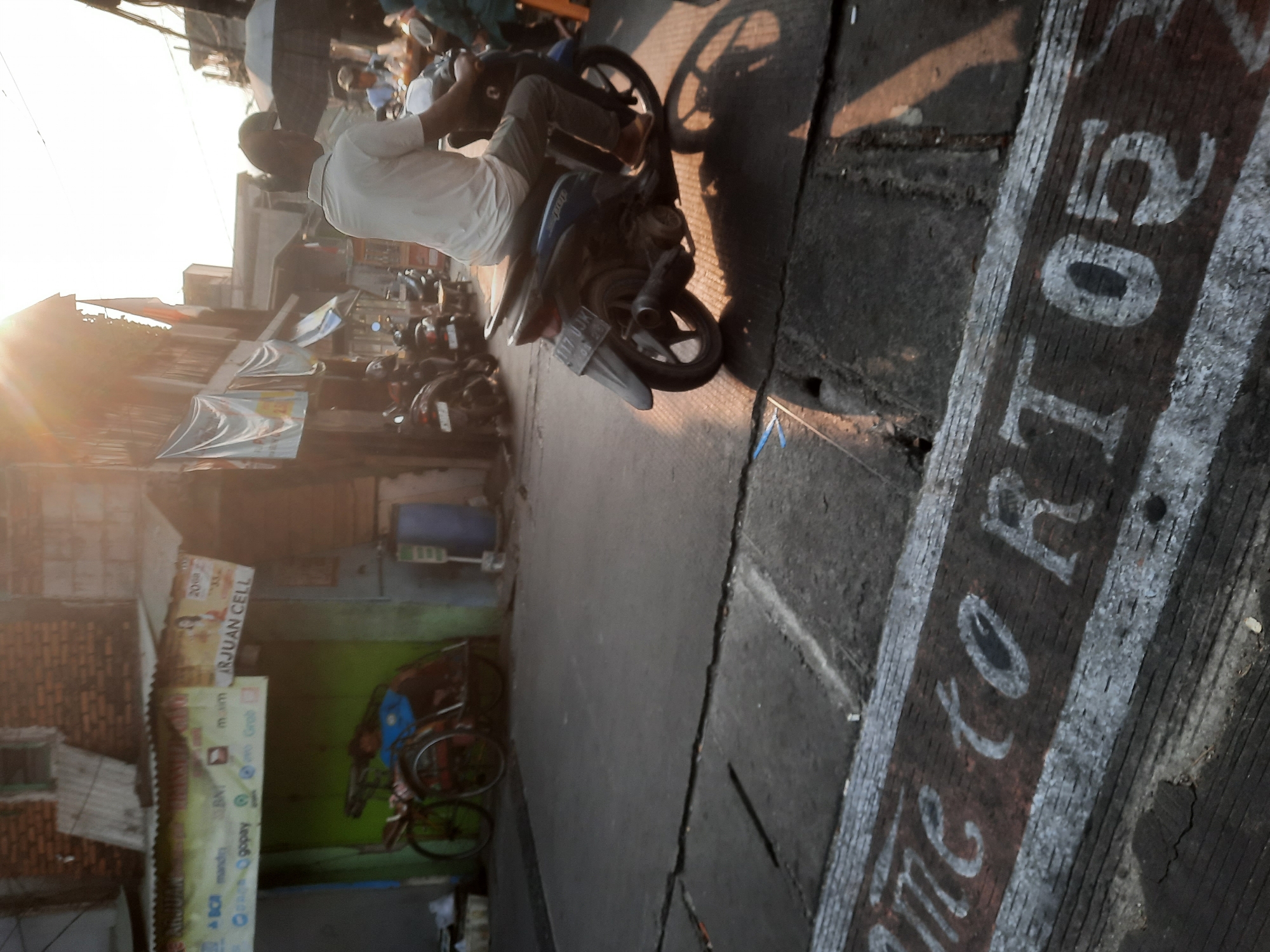 In North Jakarta, Indonesia, a person is riding a motorcycle on a cracked asphalt road during sunset. The sun is low in the sky, casting long shadows and creating lens flare. The rider, wearing a white t-shirt and grey pants, is on a blue motorcycle with a license plate that reads "B 3177 UJK". In the background, there are buildings with various signs, including one for "ARJUAN CELL," and parked motorcycles. A parked pedicab with a person inside is also visible. The ground in the foreground has painted markings that appear to spell out "MetoR 05." The overall scene depicts a street in a bustling urban environment, likely a market or residential area, during the late afternoon.