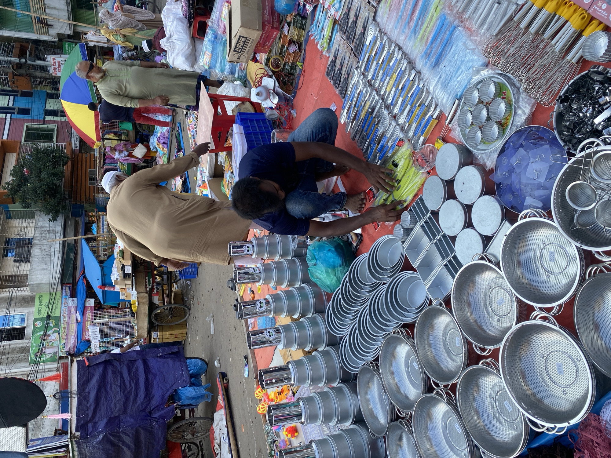 Here is a detailed description of the image:
**Scene**: A bustling street market scene in Dhaka, Bangladesh.
**People**:
* Two men are present in the market. One is kneeling and is interacting with the goods laid out on a red surface. The other man is standing, appearing to be either inspecting or bargaining for items.
**Objects**:
* A variety of kitchenware and household items are displayed on a red surface. There are rows of stainless steel bowls, strainers, cake molds, utensils, and other cooking accessories.
* In the background, there are buildings with multiple floors, vendor stalls covered with blue tarps, and various goods piled up, creating a busy market atmosphere. There is also signage visible in Bengali script.
**Environment**:
* The market is set up on a street with buildings on either side. The setting appears to be congested, with pedestrians and vendors filling the available space.