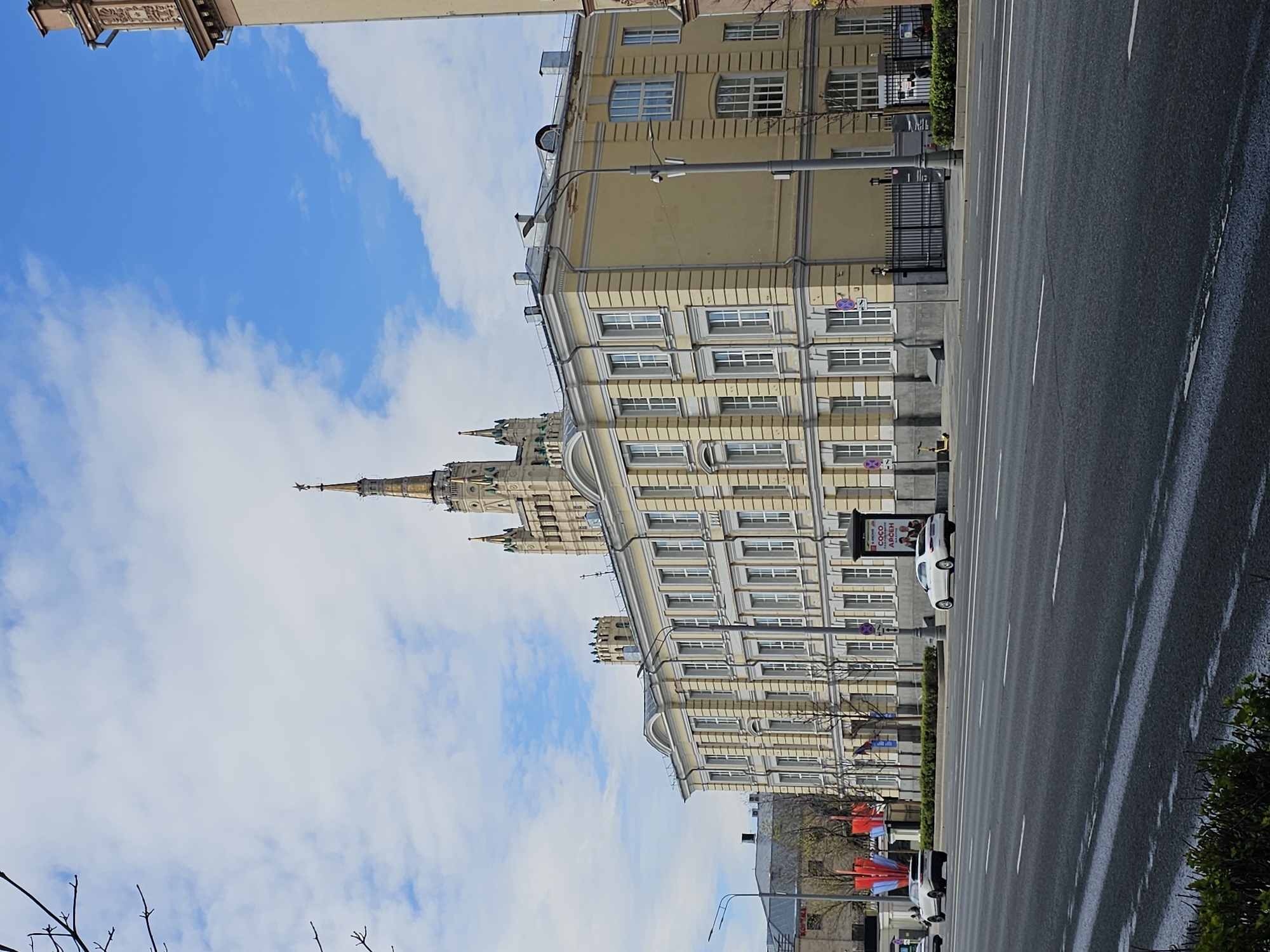 The image shows a street view in Moscow, Russia, featuring the Kotelnicheskaya Embankment Building and a portion of the Ministry of Foreign Affairs building. The foreground contains a multi-lane road with visible lane markings and vehicular traffic including a white car and other vehicles further down the street. The sidewalk is lined with greenery and advertisements. The Kotelnicheskaya Embankment Building, a Stalinist skyscraper, is visible along the right side of the image. The sky is blue with scattered clouds. Tree branches, devoid of leaves, are present in the lower left corner.