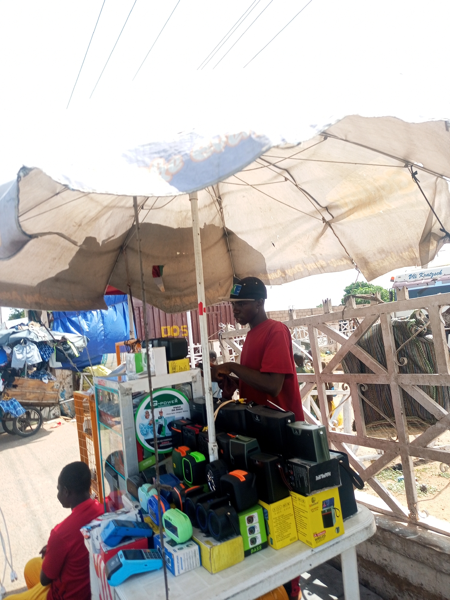 This image captures a vibrant street vendor scene in Gombe, Nigeria, bustling under the bright daylight. The main subject is a makeshift market stall set up on a street or roadside, heavily shaded by a large, worn light-colored umbrella.
Two men are visible at the stall. One man, wearing a red t-shirt and a dark baseball cap, stands behind a white table displaying various electronic gadgets. He appears to be attending to the merchandise or a customer. His profile is visible, and he looks focused on an item on the table. The second man, also in a red shirt, is seated lower down, largely obscured, with only his back and head visible, facing away from the camera towards the left.
The white table, serving as the display counter, is laden with a variety of goods, primarily electronic accessories. These include numerous portable speakers in various colors (black, green, orange, blue), power banks, and at least two bright blue and light blue Point of Sale (POS) terminals. Many items are packaged in cardboard boxes, some notably yellow. A small, clear display case is also part of the setup.
In the background, to the left, a wooden cart piled high with what appears to be clothing or fabric suggests other market activities. Behind the stall and the men, a white painted metal fence or barrier runs across the middle ground, with glimpses of dusty ground and other structures behind it. A white vehicle, possibly a truck, with "Uli Kraatzsch" written on its side, is partially visible behind the fence on the right. Another structure, possibly a container or building, is seen further back. The overall setting indicates an active outdoor market environment, likely under a sunny sky given the necessity of the large umbrella for shade.
Visible text on the products includes "3-POWER", "HAPLIFE", "BASS", and "WIRELESS", indicating types and brands of the electronic goods sold. The vehicle in the background also displays "Uli Kraatzsch".