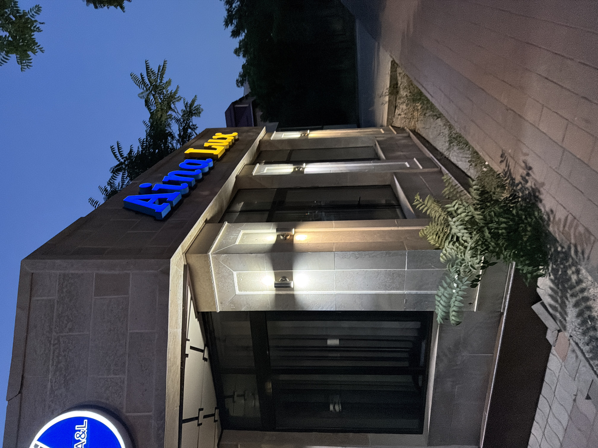 A corner view of a modern building's facade, possibly a commercial establishment, is captured at dusk. The building features a stone exterior with prominent lighting fixtures illuminating the entrance area and a portion of the facade. A large, circular sign with blue neon lettering, partially visible and reading "A&L", is mounted on the upper left corner. Above the main entrance, a larger, more prominent sign with blue and yellow neon letters spells out "Aina Lux". The building has large dark-tinted windows. In front of the building, a paved walkway is visible, leading away from the structure. A small cluster of green foliage grows near the base of the building. The background shows a street and trees silhouetted against a darkening blue sky, suggesting an urban or suburban setting in Shymkent, Kazakhstan. No people are visible in the image.