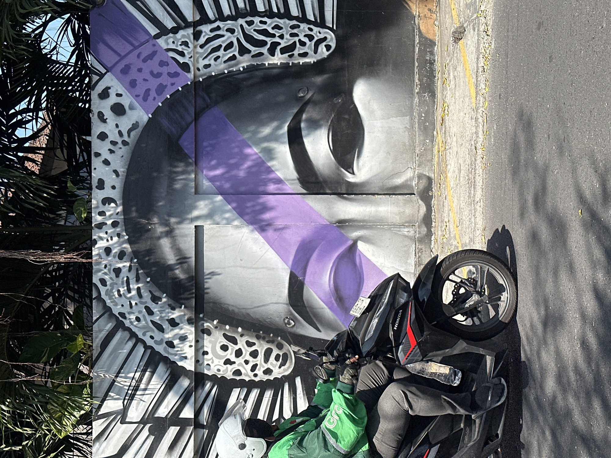 The image depicts a street scene in North Kuta, Indonesia. A person wearing a green jacket with a white helmet is riding a black motorcycle. The motorcycle has a "HONDA" logo on the front. Behind the motorcycle, there is a large mural painted on a wall, featuring a stylized face with purple accents and intricate patterns. The mural appears to be a portrait. The street is paved, and there are yellow lines painted on the side of the road. Trees with lush green leaves and hanging vines are visible above the mural, suggesting an outdoor environment with tropical vegetation. The lighting indicates it is daytime, with shadows cast on the road.