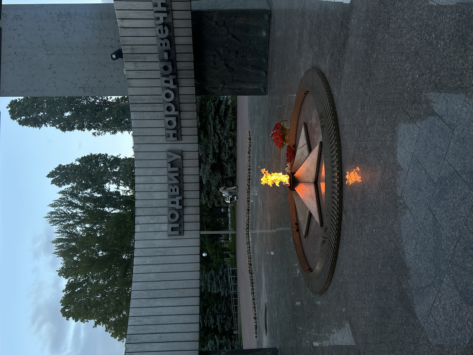 The image depicts a memorial complex, likely a war memorial, with an eternal flame burning in a five-pointed star-shaped enclosure. The structure is made of granite and metallic materials, and the inscription in Cyrillic text reads "ПОДВИГУ НАРОДА-ВЕЧНО." There are cut flowers at the base of the structure. The surrounding area has trees and a walkway leading away from the memorial. Based on location, this is the Monument of Glory in Tolyatti, Russia.