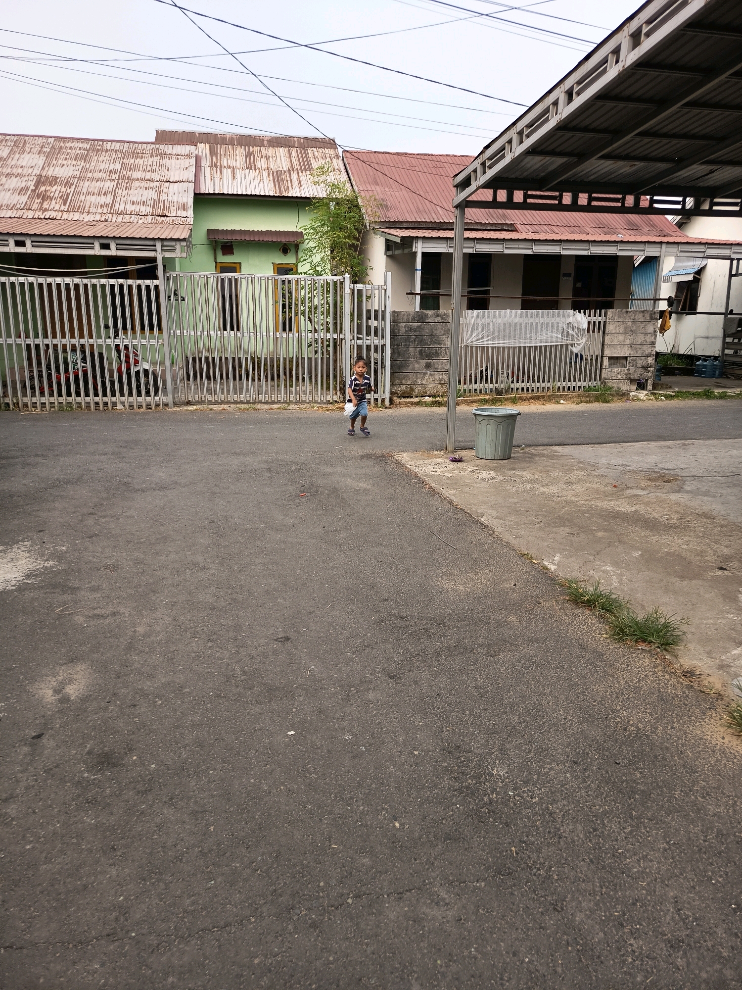 A young boy wearing a striped t-shirt and shorts is running on an asphalt road in front of several houses. He is holding a white object in his right hand. The houses have metal fences in front of them and corrugated metal roofs. A green house with yellow window frames is visible on the left. A covered parking area with a metal roof is on the right. A grey trash can sits on a concrete path next to the road. The scene appears to be in an urban or suburban area of Banjarmasin, Indonesia, with overhead electrical wires visible against a cloudy sky.
