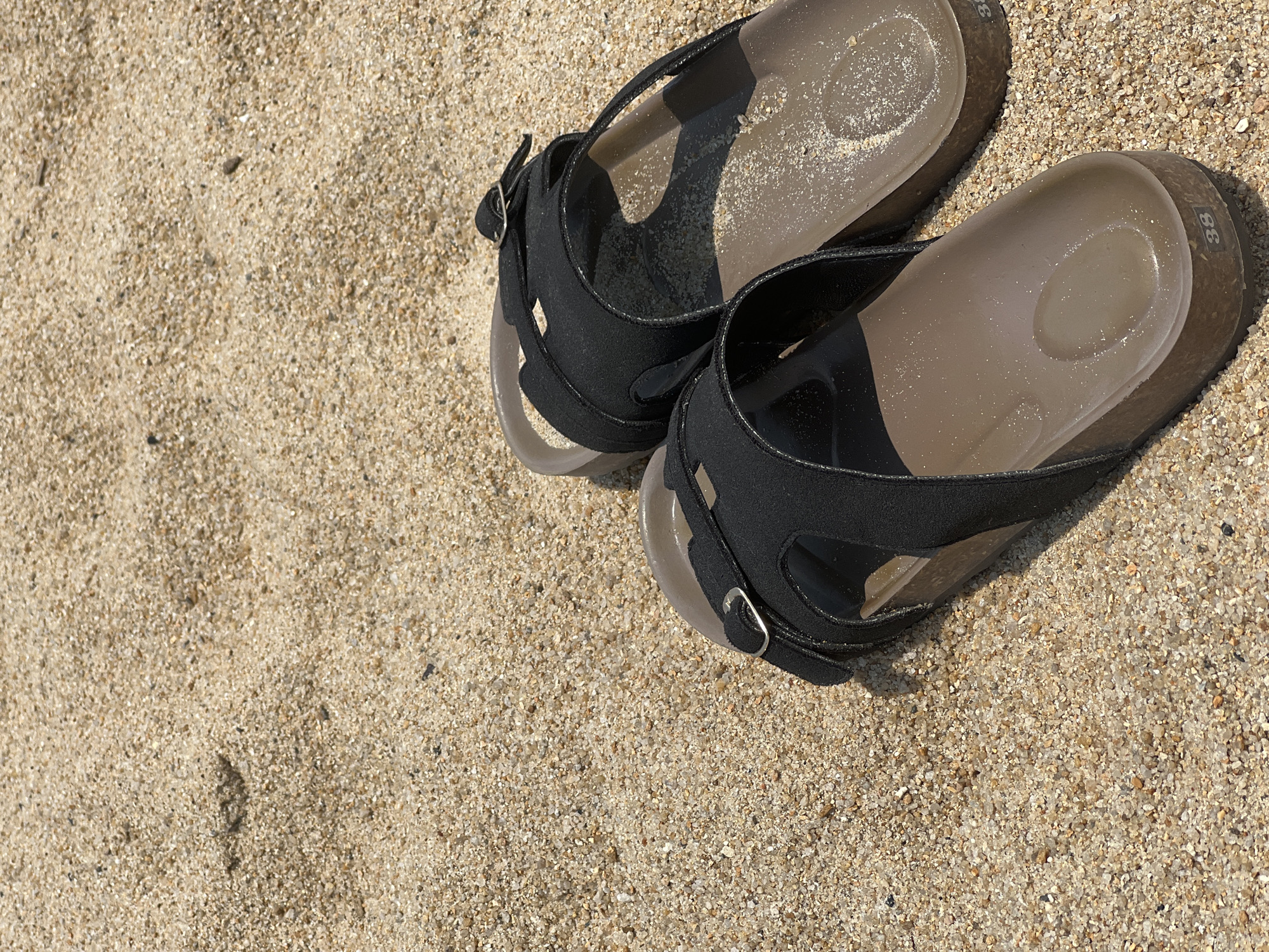 The image depicts a pair of women's black sandals resting on light brown sand. The sandals are size 38. The sand appears coarse. The location is consistent with a beach environment.