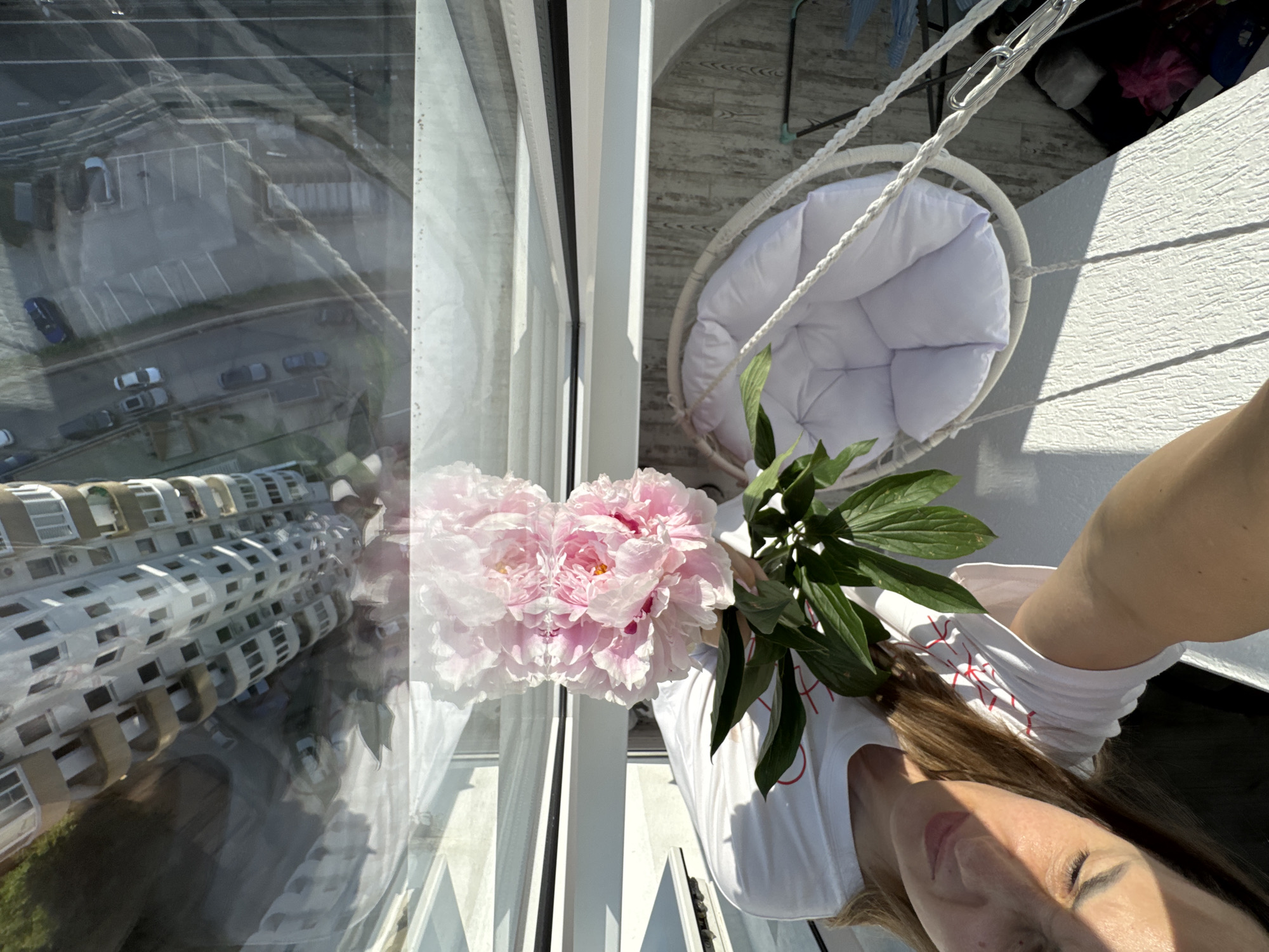 A woman is taking a selfie on a balcony in Sochi, Russia, holding a bouquet of pink peonies. The balcony overlooks a parking lot and a multistory building. She is wearing a white t-shirt with red lettering and her hair is brown. A white hanging chair is visible on the balcony. The image is taken from a high angle, looking down.