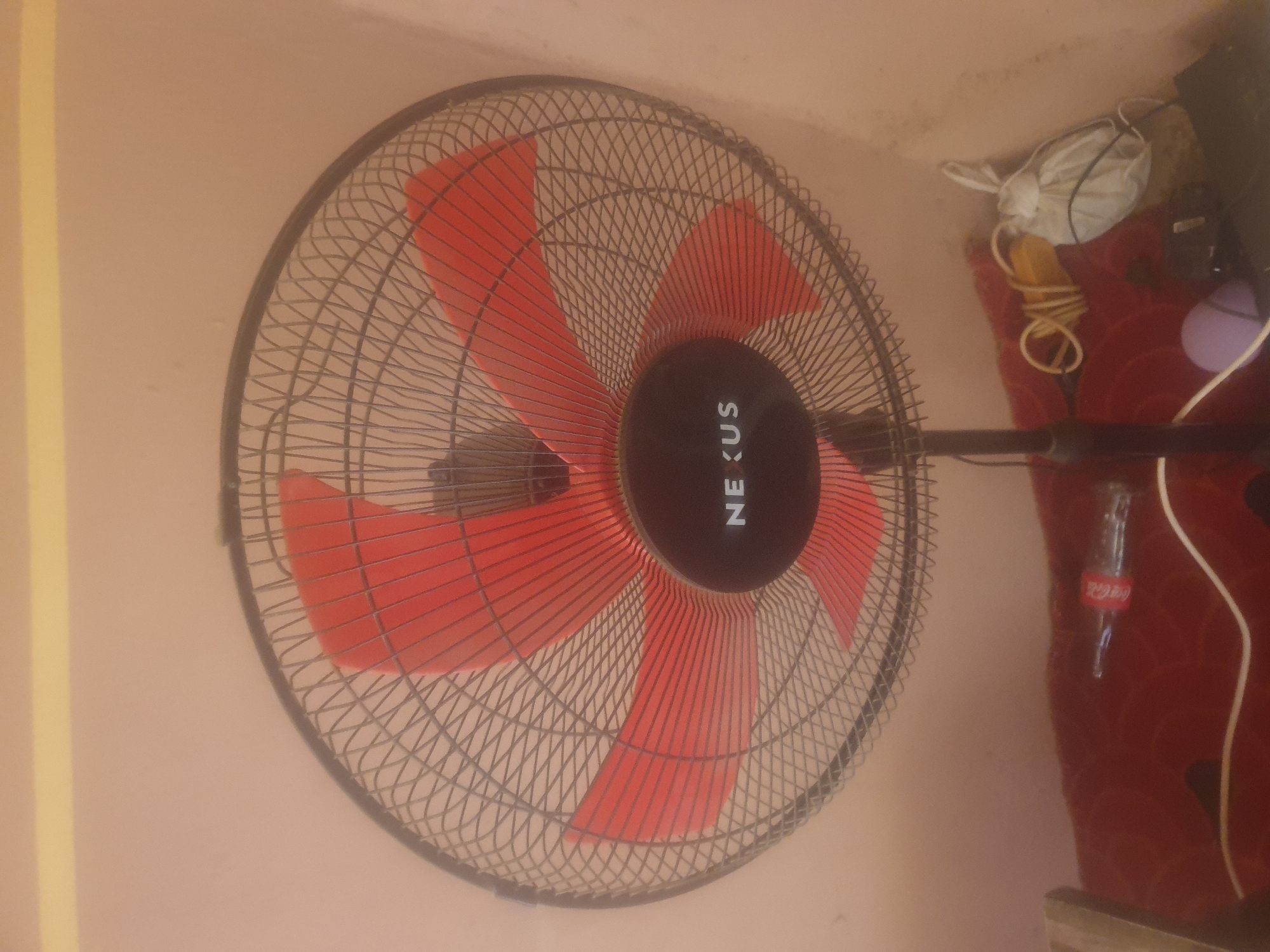 An electric fan with red blades and a black grill sits on a stand. The fan has "NEXUS" written in white letters on a black circular housing in the center. The fan is positioned in front of a light pink wall, and below it, on a red surface, is a bottle of Coca-Cola, a coiled white cord, a small yellow object, and a white plastic bag. The image appears to be taken indoors, likely in a residential setting. There is no indication of the city of Washington or the United States of America.