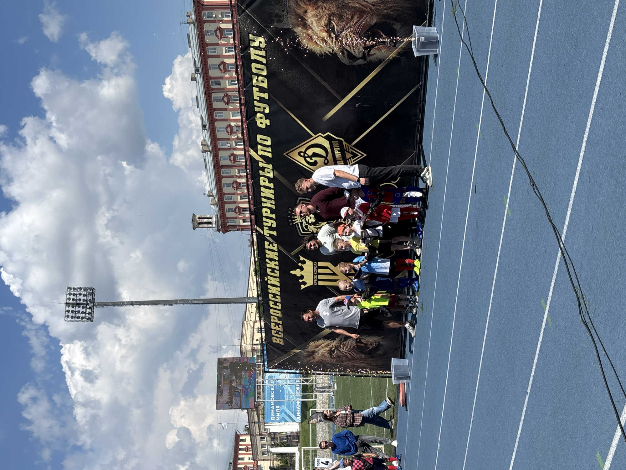The image depicts an outdoor event, likely a children's football tournament, taking place on a blue running track in Izhevsk, Russia. A large black banner with golden lettering, reading "ВСЕРОССИЙСКИЕ ТУРНИРЫ ПО ФУТБОЛУ" (All-Russian Football Tournaments) and featuring a prominent image of a lion's head and a logo, serves as the backdrop. Several adults and children are gathered in front of the banner. The adults appear to be organizers or coaches, while the children are dressed in football uniforms. Some of the children are holding a black banner or ribbon, and there are celebratory sparks or confetti visible in the air. In the background, there are stadium lights, buildings, and a blue sky with clouds. The event seems to be well-attended and festive.