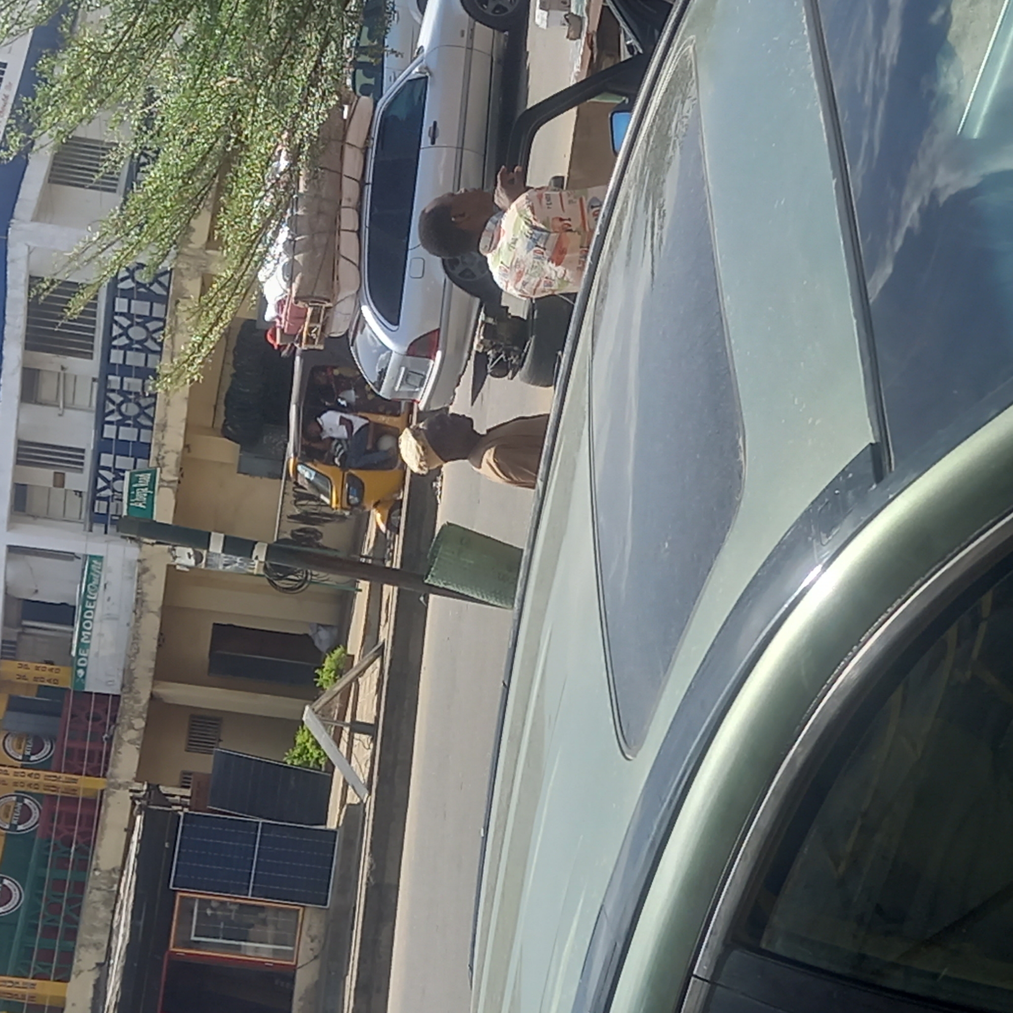 The image depicts a street scene in Dadi Riba, Nigeria, captured from inside a vehicle, focusing on a street. There's a building with signage and commercial advertisements on the left. The street has other vehicles, a yellow auto rickshaw (a type of motorized tricycle), and a few pedestrians. Two people are conversing.