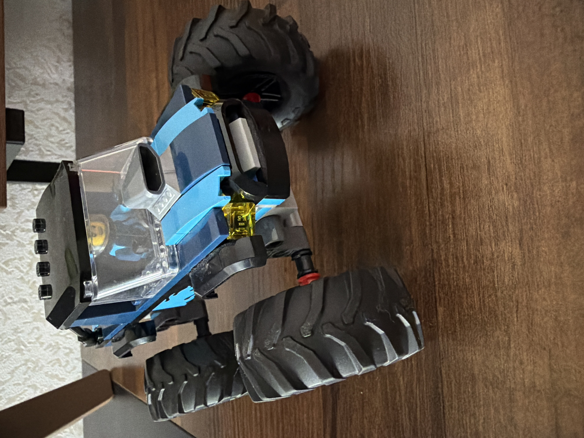 A toy monster truck, primarily blue and black with clear plastic windows, is positioned on a wooden surface. The truck has large, knobby tires. The scene appears to be indoors, possibly on a table or floor with a wood grain pattern. There are no people visible. The lighting suggests it could be daytime, but no specific time of day can be determined. No weather conditions are apparent. The location context is Yablonovskiy, Russia, but this is not visually evident in the image. There is a small yellow label with black text on the truck, but the text is too small and blurry to be read.
