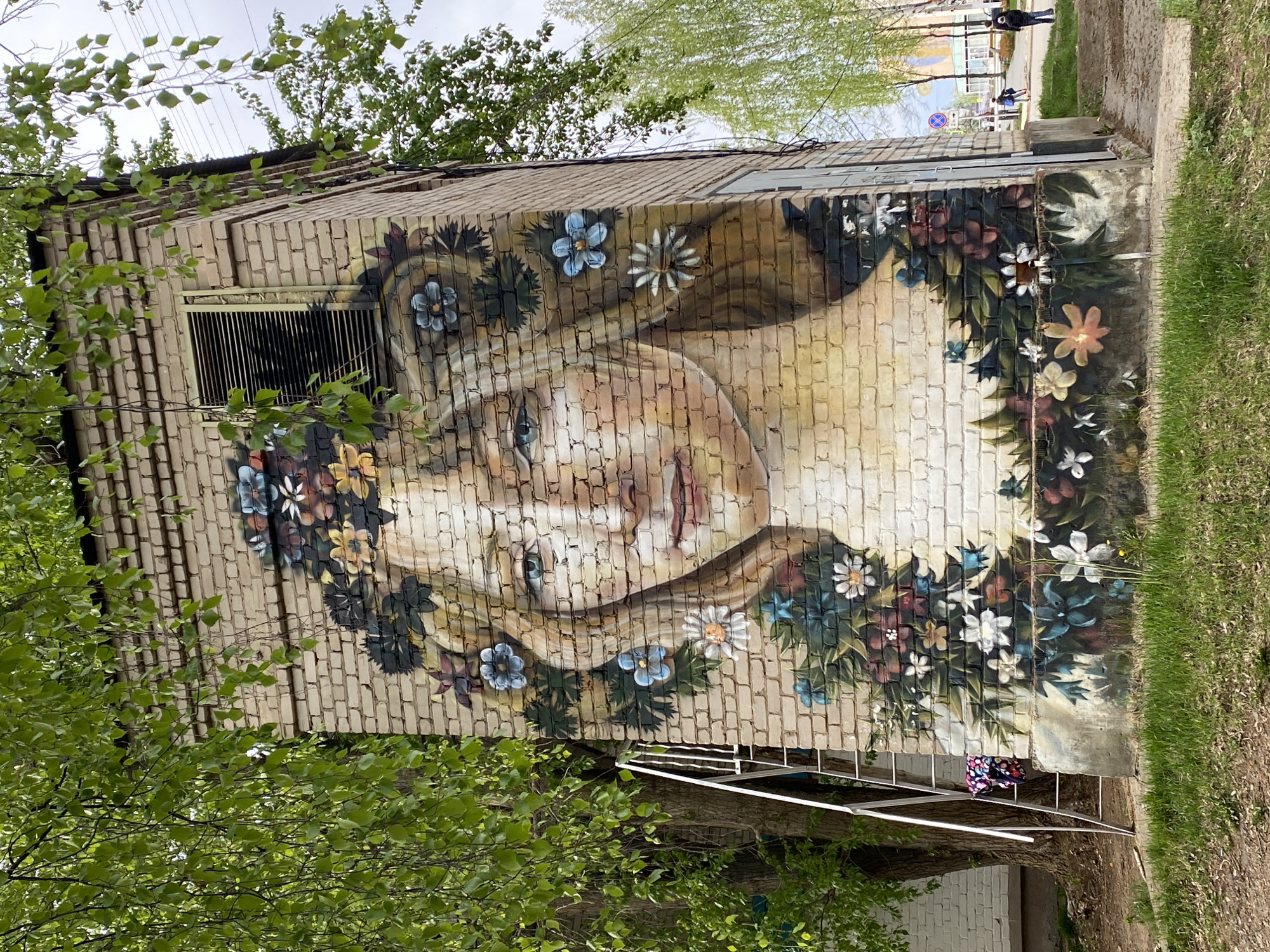 A brick building in Chaikovsky, Russia, features a mural of a woman's face adorned with flowers. A metal staircase runs along the building's side. Trees with green foliage flank the building. People are visible walking along a path in the background. Patches of grass surround the building.