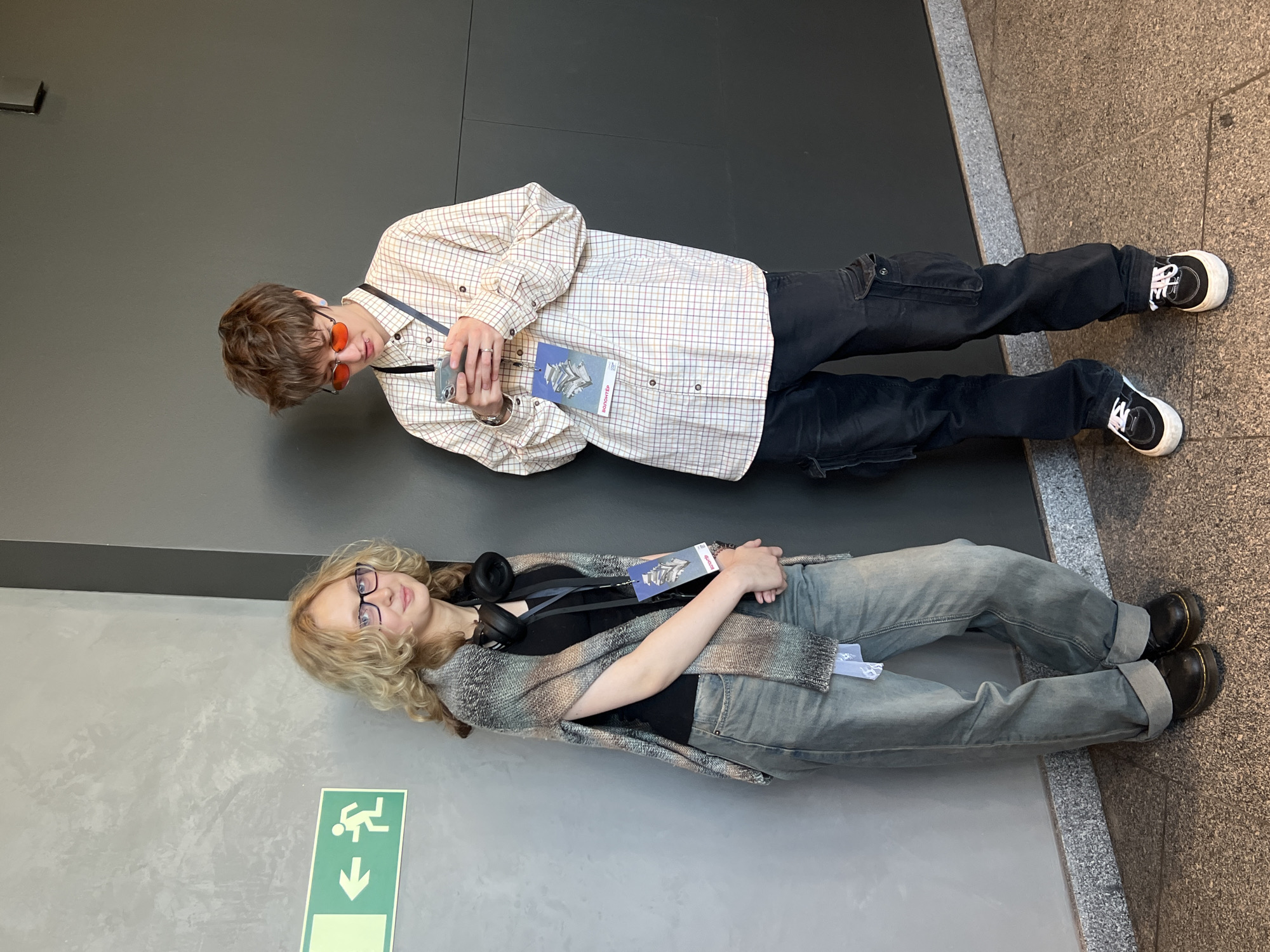 Two young adults are lying on the floor next to walls in a brightly lit interior space.
The first individual is male, has short brown hair, and wears small orange sunglasses, a checkered button-down shirt, black cargo pants, and black and white sneakers. He is holding a smartphone and wearing an identification badge around his neck.
The second individual is female, has blonde wavy hair, and wears glasses, a black t-shirt, a patterned knitted shawl, light-wash blue jeans, and black Doc Martens boots. She also has headphones around her neck and wears an identification badge.
The walls are different shades of gray. There is a green and white emergency exit sign on the wall next to the female. The floor is made of light gray stone tiles.
The badge has the word "BOTANIKA" printed on it, a brand of Saint Petersburg luxury hotel. This suggests that the image was taken somewhere in that city.
