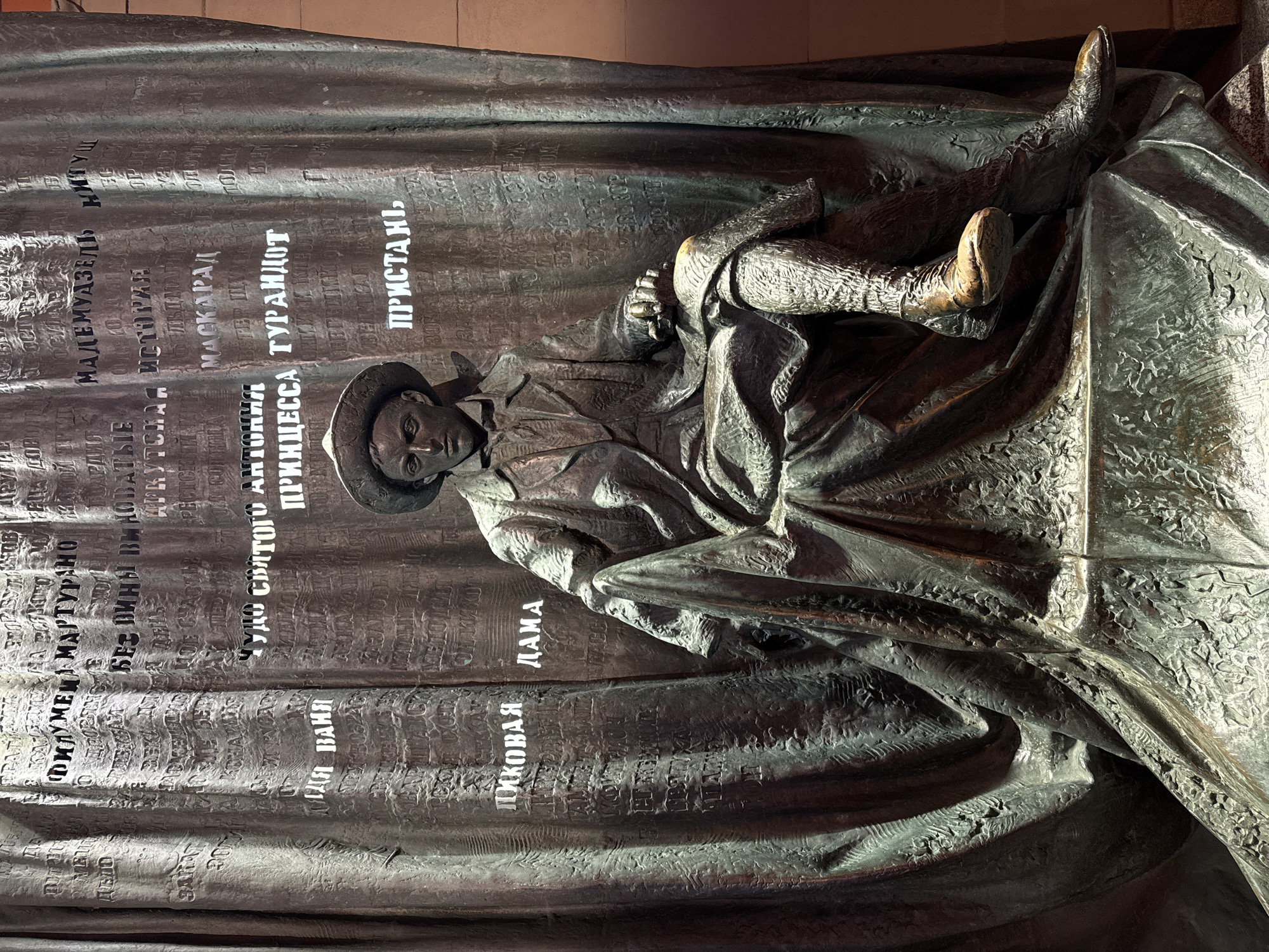 The image shows a bronze sculpture depicting a man lying down in what appears to be a dramatic pose, possibly representing death or sleep. The sculpture is attached to a textured metal surface with raised lettering. The Russian text includes theatrical references, such as "Dama" (Lady), "Pikovaya" (of Pique), "Turandot," and "Dyadya Vanya" (Uncle Vanya). The sculpture is part of a memorial theatre sculpture complex in Moscow, Russia.