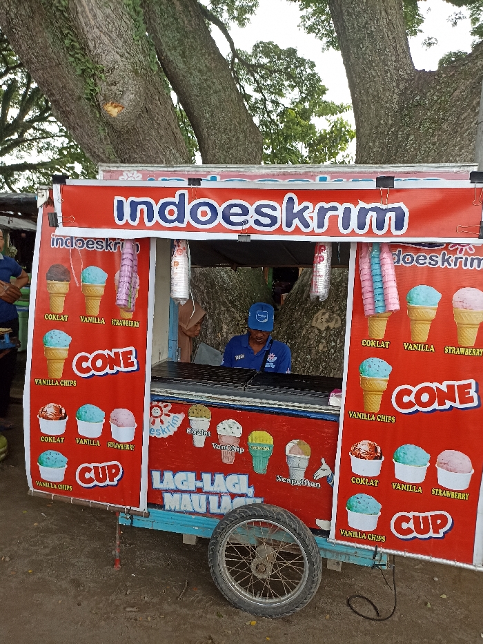 An outdoor scene captures an "indoeskrim" ice cream vendor and his cart, likely in Kepung, Indonesia. A male vendor, wearing a blue cap and shirt, is visible behind the counter of the bright red and blue cart, ready to serve. Another person is partially visible on the far left.
The ice cream cart is the central focus, adorned with colorful images of various ice cream cones and cups. Hanging from the cart's canopy are stacks of colorful ice cream cones (pink, blue) and plastic cups, presumably for serving. A single spoked wheel of the cart rests on the dirt ground at the bottom right.
The setting appears to be a casual public area, possibly a street or park, characterized by the massive trunks and green foliage of a large, mature tree providing natural shade in the background. The lighting suggests it is daytime.
The cart's signage prominently displays "indoeskrim" multiple times. It advertises ice cream flavors including "COKLAT" (Chocolate), "VANILLA," "STRAWBERRY," and "VANILLA CHIPS," available in both "CONE" and "CUP" formats. Other discernible text includes "Scapolitan," "eskrim," and the Indonesian phrase "LAGI-LAGI MAU LAGI," which translates to "Again and again, want more again," a catchy marketing slogan. The scene portrays a typical local street food operation.