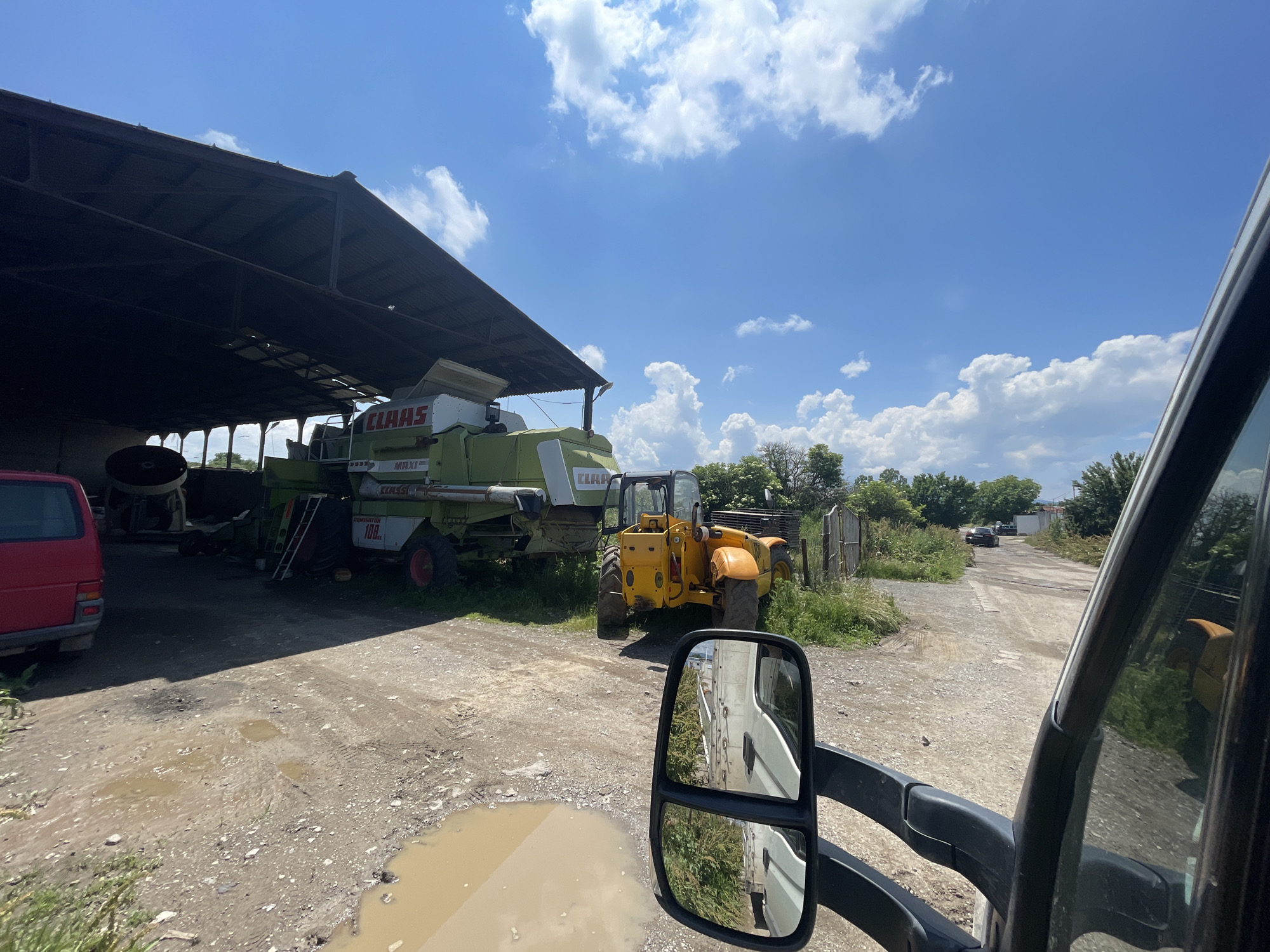 The image presents a daytime scene at what appears to be a farmyard or industrial storage area in Gorni Bogrov, Bulgaria. The perspective is from inside a vehicle, likely a van or truck, with a large black side mirror and part of the window frame occupying the right side of the frame.
In the midground, under a large, open-sided shed with a dark roof, a green combine harvester is prominently parked. Visible text on the combine includes "CLAAS", "MAXI", "108", and "SE". To its left, a red van is partially visible. Next to the combine harvester, a yellow telehandler is parked on a dirt and gravel surface, near a wooden fence and some overgrown grass.
The ground is unpaved, showing tire tracks and several muddy puddles that reflect the bright sky. Beyond the machinery and fence, a dirt track or road stretches into the background, flanked by lush green trees and bushes. A dark-colored car can be seen further down this track.
The sky is a vibrant blue with scattered white, cumulus clouds, indicating a bright and sunny day. No people are visible, and the scene depicts parked vehicles and machinery rather than active work.