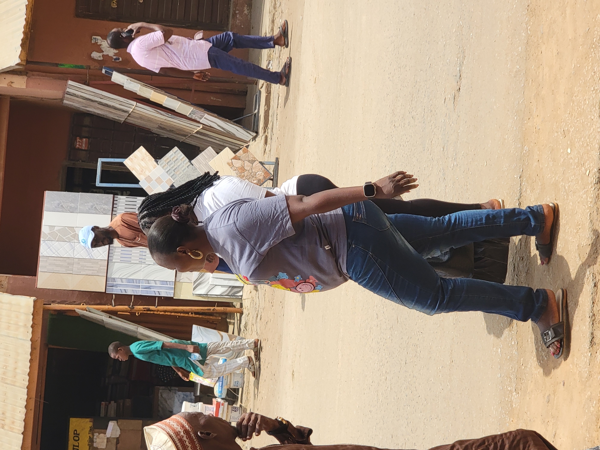 The image captures a bright, sunny street scene in Maiburiji, Nigeria, with multiple individuals and a prominent display of building materials.
In the mid-ground, a person wearing a grey t-shirt with a colorful graphic, blue jeans, and brown slide sandals stands, their left arm slightly bent. They have braided hair, a gold hoop earring, and a dark smartwatch on their left wrist. Behind them, another person with dark hair, in a white top and dark pants, is partially visible. Further back, to the left, a person in a light blue cap and brown shirt is seen. On the right side, a man in a pink checkered shirt and blue pants holds a phone to his ear, seemingly in a conversation. In the lower foreground, part of a person's head, wearing a patterned cap (possibly a kufi) and brown clothing, is visible. To the left, another individual in a green shirt and patterned trousers stands near an open shop entrance, holding a bottle.
The setting is an outdoor commercial area, likely a market or street lined with shops, given the various stacks of patterned and plain ceramic tiles displayed. The ground is unpaved, appearing sandy or dusty. Buildings with reddish-brown walls form the background. The bright daylight casts distinct shadows, indicating a clear, sunny day. On a yellow sign in the lower left, the partial text "LOP" is visible. The scene depicts a typical day of activity and commerce in Maiburiji.