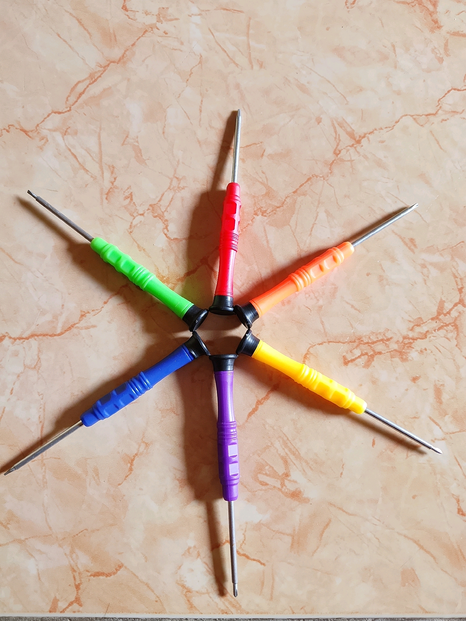A top-down, eye-level, medium shot captures a set of six precision screwdrivers arranged in a star pattern on a light brown, marble-patterned tiled surface. The screwdrivers, each with a different colored handle - red, orange, yellow, purple, blue, and green - radiate outwards from a central point where their black connector bases meet. The metallic shafts of the screwdrivers extend towards the edges of the frame, with their pointed tips visible. The lighting is natural and overhead, casting subtle shadows from the screwdrivers onto the tiled surface. There are no people visible in the image. The scene is clean and organized, suggesting a tool kit or a workspace. The marble-patterned tile is a common flooring material found in residential and commercial spaces. This image was captured in Pelaihari, Indonesia.
