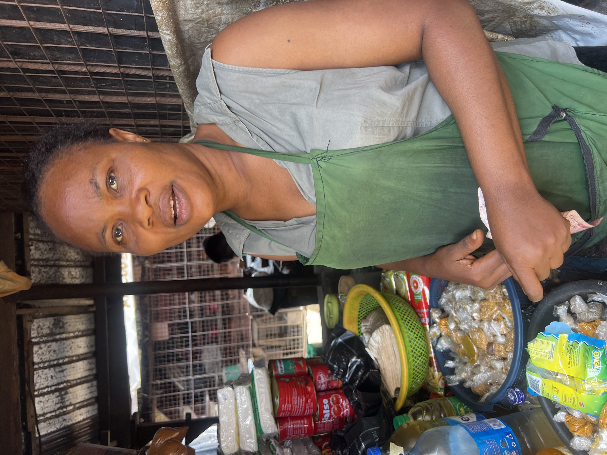 A woman stands behind a market stall in Nkpor, Nigeria, wearing a green apron over a grey sleeveless shirt. She is looking slightly off-camera with her mouth open, as if speaking. The stall is stocked with various goods, including cans of tomatoes, packets of tissue, bottles of oil, and baskets filled with small, individually wrapped food items. In the background, a metal grate structure suggests the market setting. The overall scene depicts a typical day at a local Nigerian market.
