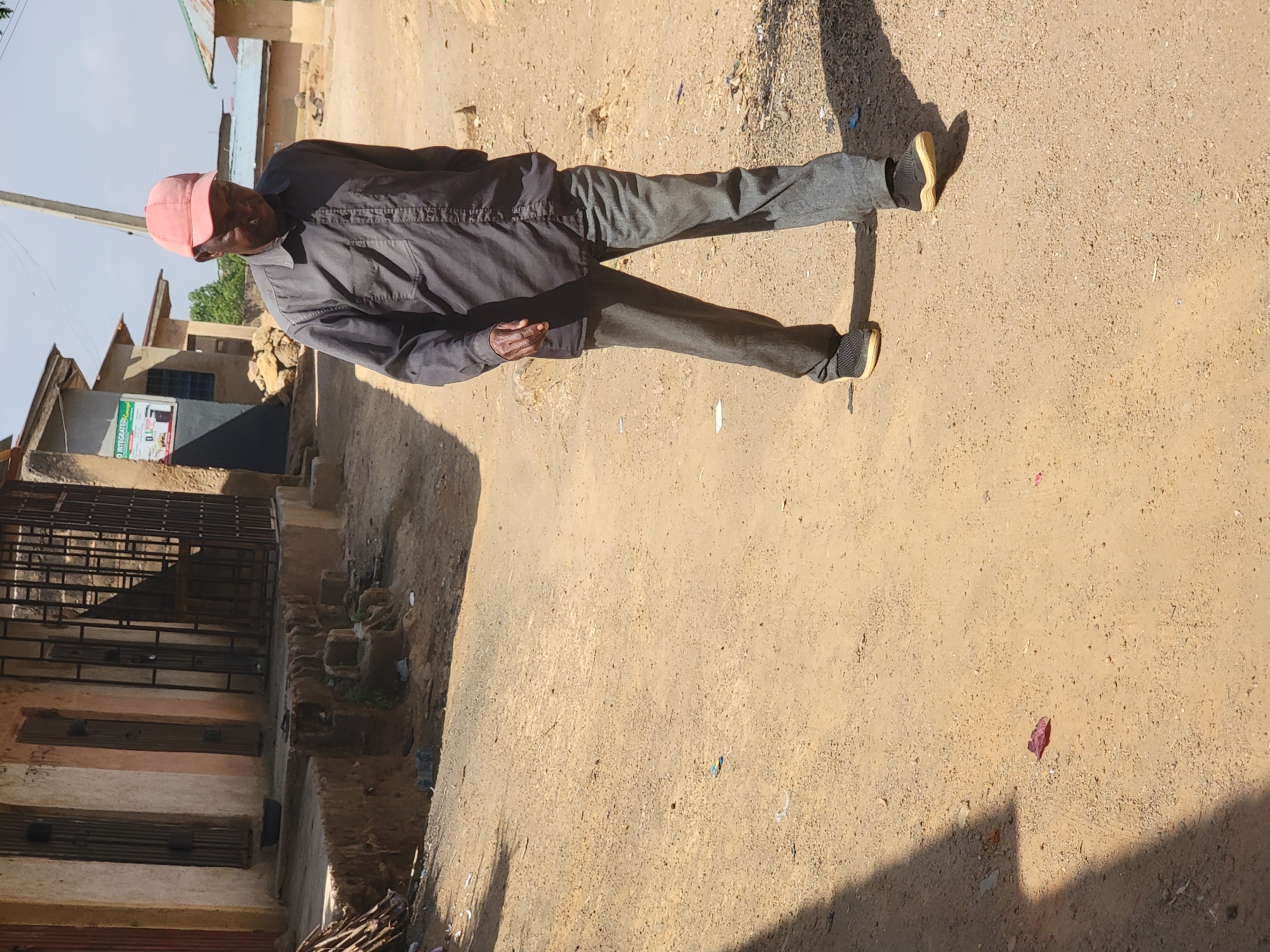 The image shows an older man walking on a dirt street in what appears to be a residential area. He is wearing a pink cap, dark-colored shirt, and gray pants with sneakers. The location is possibly Jos, Nigeria, as requested. The background shows buildings with metal gates and a sign with some writing on it. The ground is sandy and there is some trash scattered around. The light suggests it is daytime, and the shadows indicate a strong source of sunlight.