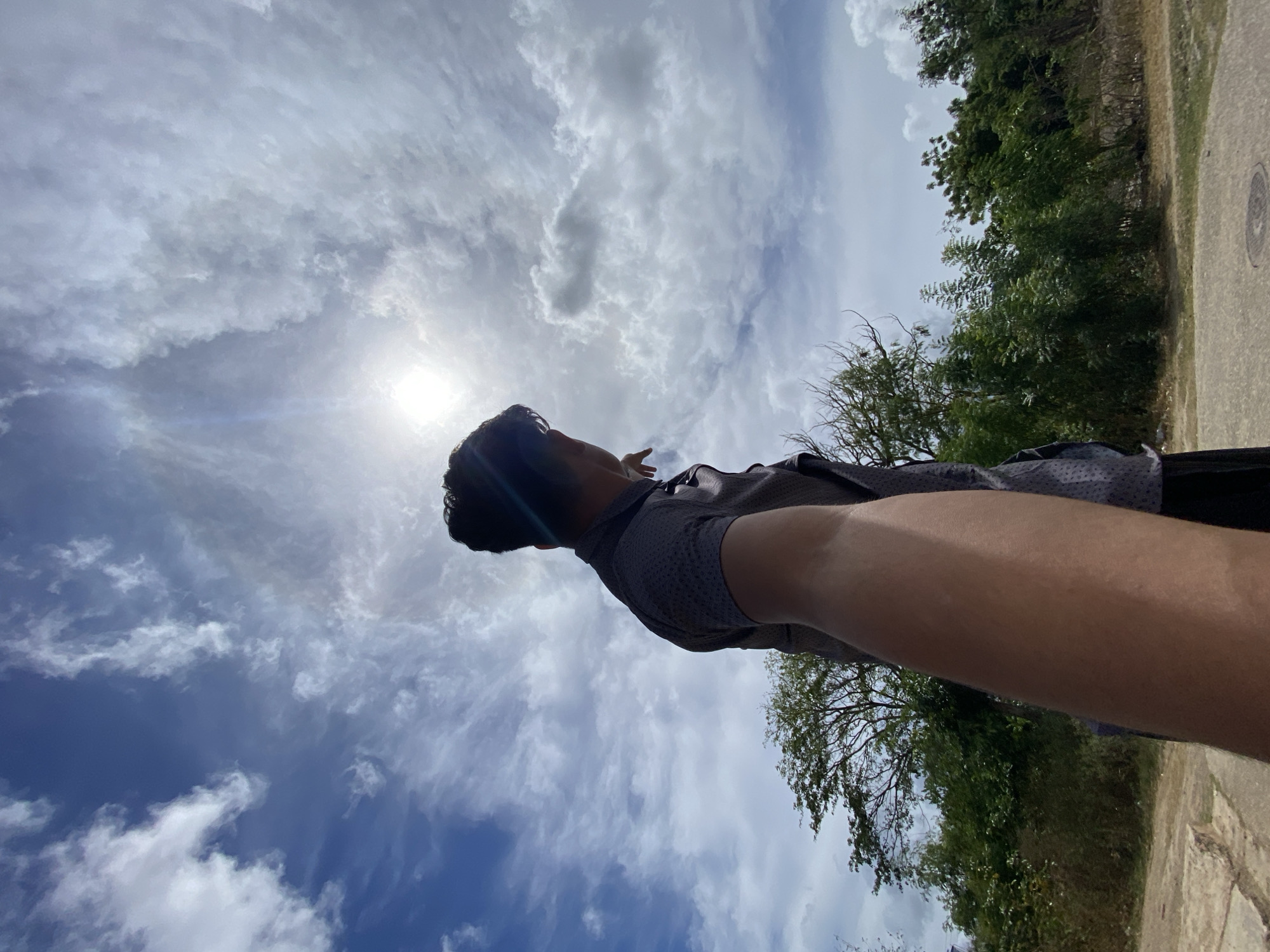 An outdoor scene in Juan Griego, Venezuela, depicts a person from a low-angle perspective, captured from behind and slightly to the right. The individual, dressed in a dark-colored, short-sleeved patterned shirt, faces towards the upper left of the frame. Their right arm is extended directly towards the camera, with the index finger of their right hand pointing upwards and slightly left towards the bright sun, which is prominent in the upper left quadrant of the sky and creates a lens flare effect. The sky occupies approximately two-thirds of the image and features a mix of blue patches and scattered white clouds. The lower third of the image shows ground cover, consisting of a light-colored, possibly concrete or paved surface on the left and a darker, unpaved area on the right. Dense green foliage, including trees and shrubs, forms the background, suggesting a natural or semi-natural environment.