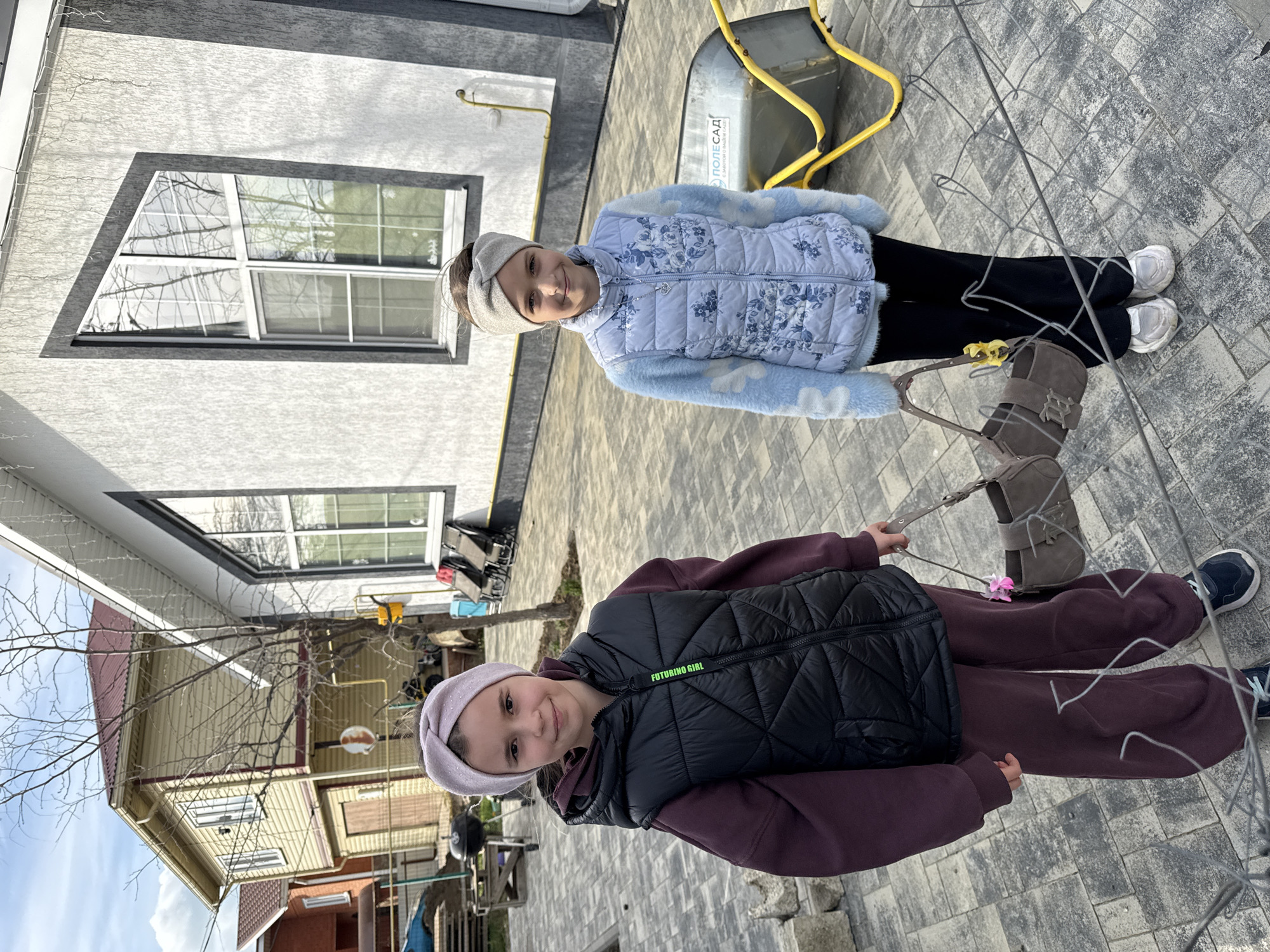 Two young girls stand on a gray brick-paved area. One wears a light blue floral vest, black pants, and white sneakers, carrying a gray purse. The other wears a black vest over a dark purple tracksuit, holding the purse strap. A wire fence is in the foreground. Behind them are buildings with varied architectural designs. A yellow object resembling a snow pusher is also visible. The location appears to be an urban or suburban setting. While not explicitly stated, given the request, the location is presumed to be Taman, Russia.
