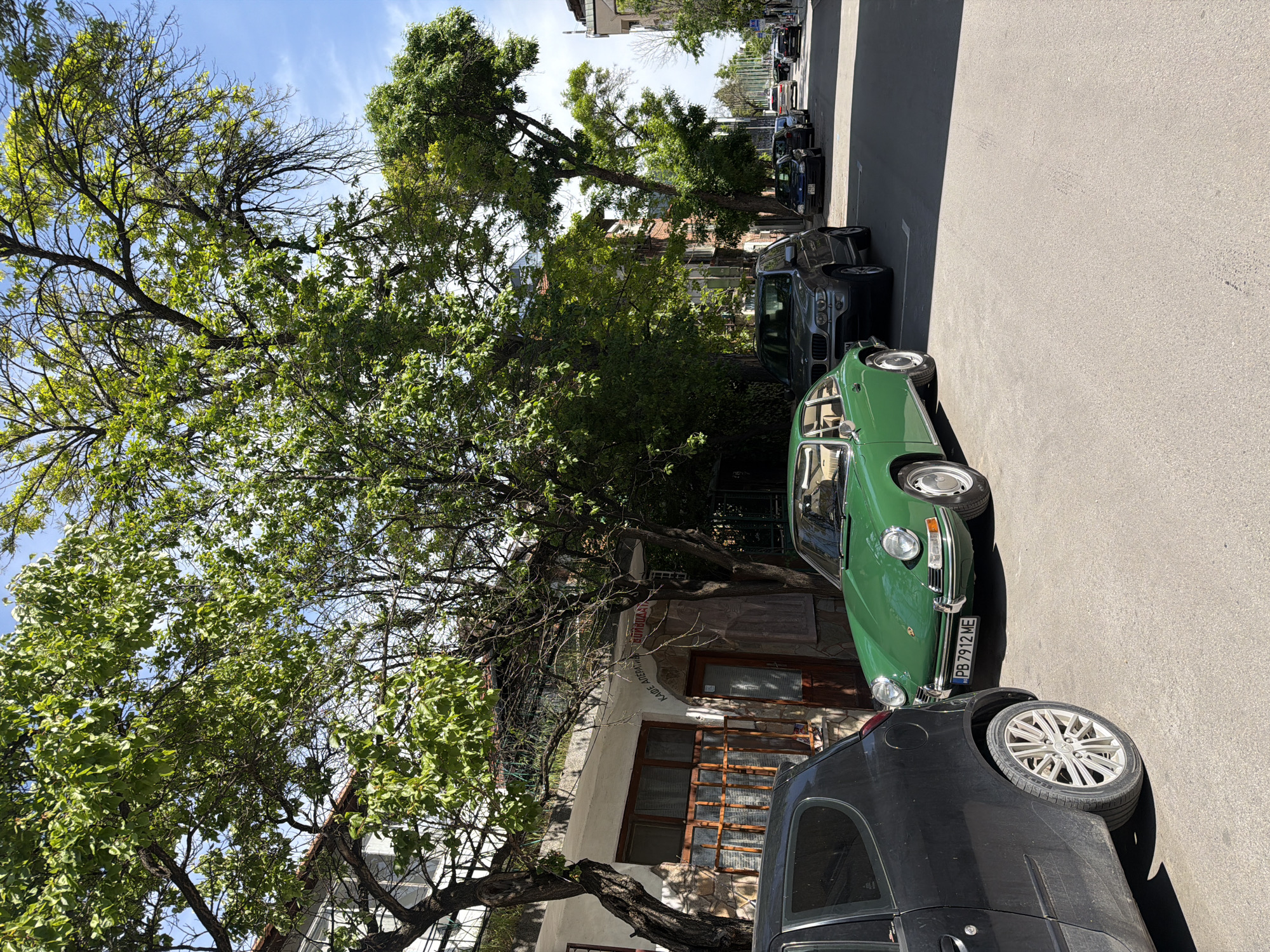 The image shows a street scene in an unidentified city. The view is from street level, looking along a line of parked cars. The first car is black, followed by a green classic car and then other cars further down the street. The background is comprised of trees, buildings, and a bright blue sky. The photograph is taken during daylight, casting harsh shadows on the street. The trees on the left are mature, providing a dense canopy above the street. The building visible behind the trees seems older, with wooden window frames.