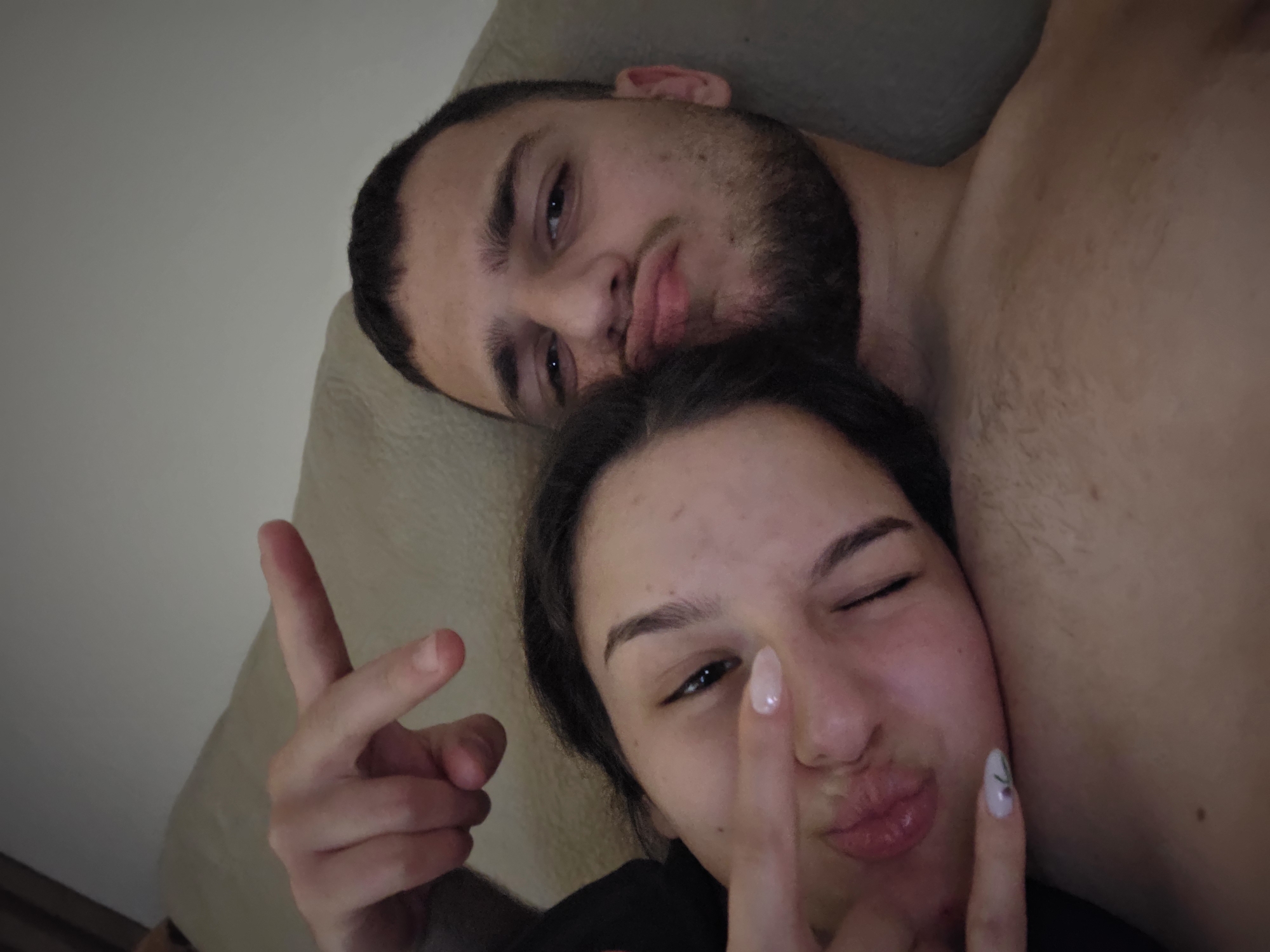 A close-up shot depicts a couple indoors. A bearded man lies on his back, with a woman positioned partially on top of him. Both are making faces, with the woman winking and holding up two fingers in a V-sign. The man also has his lips pursed. The setting appears to be a casual, intimate environment. No information given indicates the location of Stara Zagora, Bulgaria.