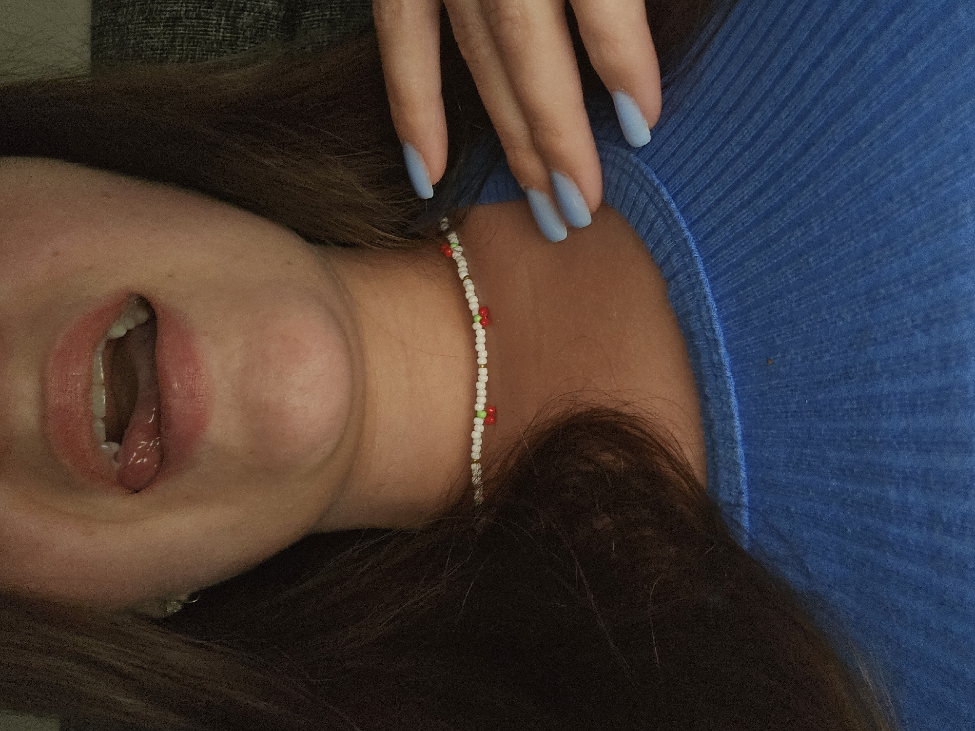 This is a close-up, waist-up shot of a young adult with long, dark brown hair, wearing a bright blue ribbed knit shirt. They are looking down and to the left, with their mouth open and tongue sticking out slightly. Their lips are glossy, and their teeth are visible. A delicate beaded necklace with white, green, and red beads, resembling cherries, is worn around their neck. To the right, their hand, with light blue painted nails, is resting on their collarbone. The background is a dark, textured surface, possibly a couch. The scene is indoors and dimly lit. There is no indication of the specific location in Petropavlivska Borshchahivka, Ukraine.