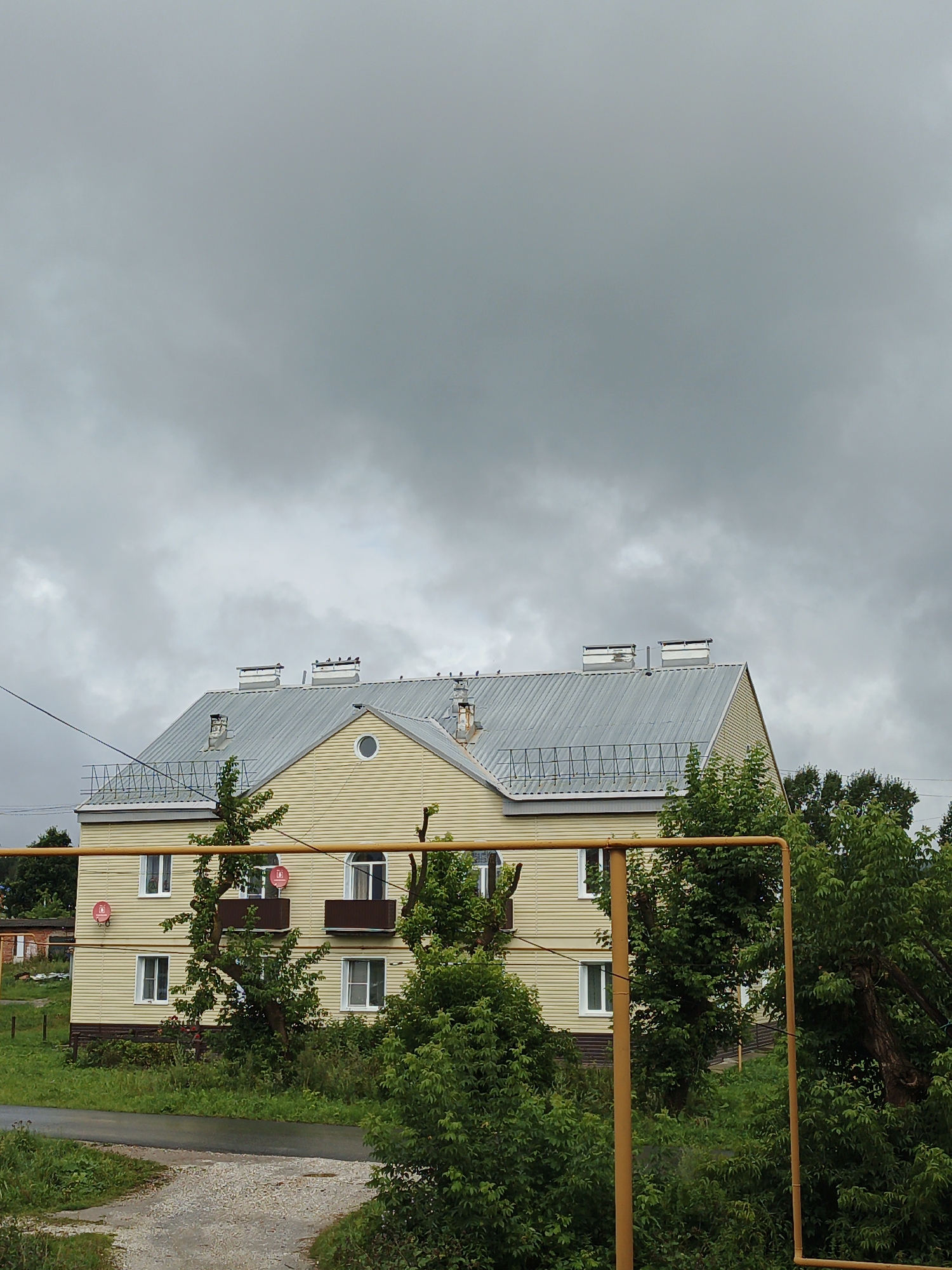 The image depicts a three-story, light yellow apartment building situated in Medvedyovka, Russia, under a cloudy, overcast sky. The building has a gray, corrugated metal roof with several ventilation stacks and what appear to be satellite dishes on the balconies of the second floor. Small trees and bushes surround the building, with a gravel and asphalt path leading towards it. A yellow metal pipe fence runs across the foreground. The scene appears to be devoid of people or any discernible activity.
