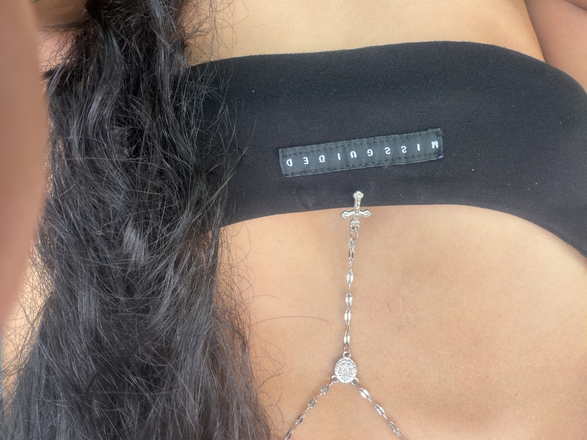 The image is a close-up shot of a person's upper torso, focusing on the chest area. The subject is wearing a black garment, likely a bandeau top or bra, with a small white tag that reads "MISGUIDED" in uppercase letters. Attached to the garment is a delicate silver body chain with a crucifix charm at the top, a circular charm with an embossed design below it, and a series of small links forming the chain. The subject has dark, wavy hair that cascades down over the left side of the frame, partially obscuring the garment and skin. The skin tone appears to be light to medium. The lighting suggests it might be daytime, with soft shadows. The setting appears to be casual, and the focus is on the fashion accessories and clothing. There are no strong indicators of emotion or a specific event. The visible text is "MISGUIDED" on the garment's tag. The location context is Baan Bang Rak, Thailand.