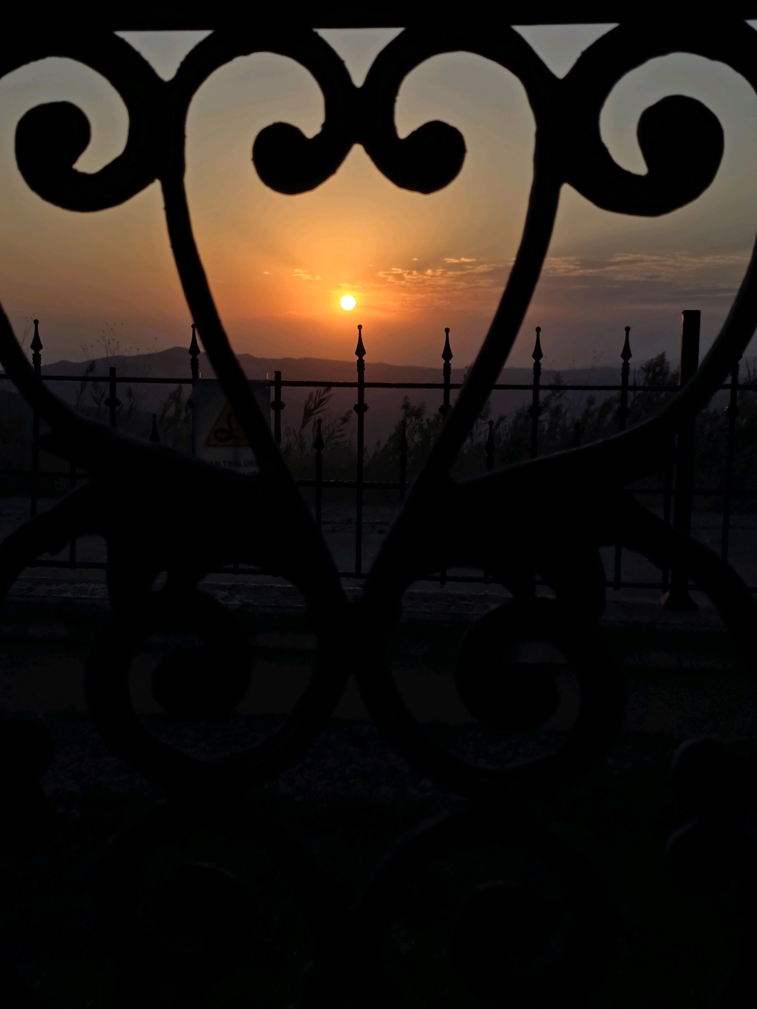The image shows a sunset viewed through an ornate metal gate. The gate has a decorative, heart-like shape in the center of the frame, with swirling patterns on either side. The sun is a bright, orange disc positioned in the upper middle of the frame, slightly to the left of center, with a few wispy clouds around it. In the background, there are rolling hills or mountains, a fence with pointed finials, and some sparse vegetation. There is a yellow hazard sign visible through the fence, with some text on it. The foreground is dominated by the dark silhouette of the gate. The location is identified as Muğanlı, Azerbaijan. The image is captured at sunset, creating a warm, orange and yellow color palette in the sky.