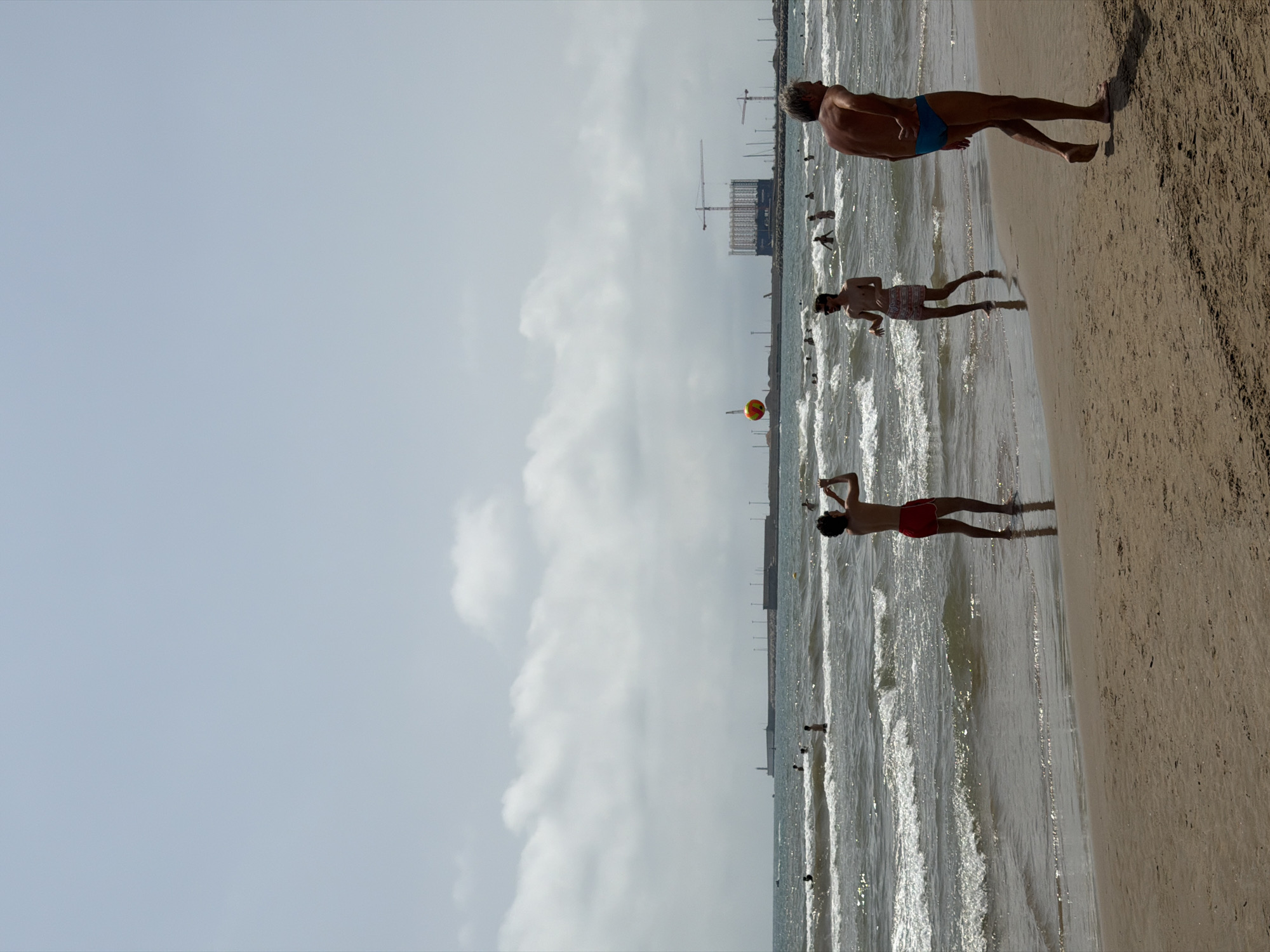 In an outdoor, daytime scene on a sandy beach, several people are present. In the foreground, a man with gray hair, wearing blue swim trunks, walks away from the viewer. Further into the scene, a young man with dark curly hair, wearing red swim trunks, stands in the shallow water and holds his hands to his face as if taking a picture. To his left, another young man in patterned swim trunks walks through the water. In the middle ground, several other people are visible in the ocean. In the background, a breakwater with lights and a lighthouse extends into the sea. Beyond the breakwater, a large building under construction with cranes is visible. The sky is overcast with light clouds.