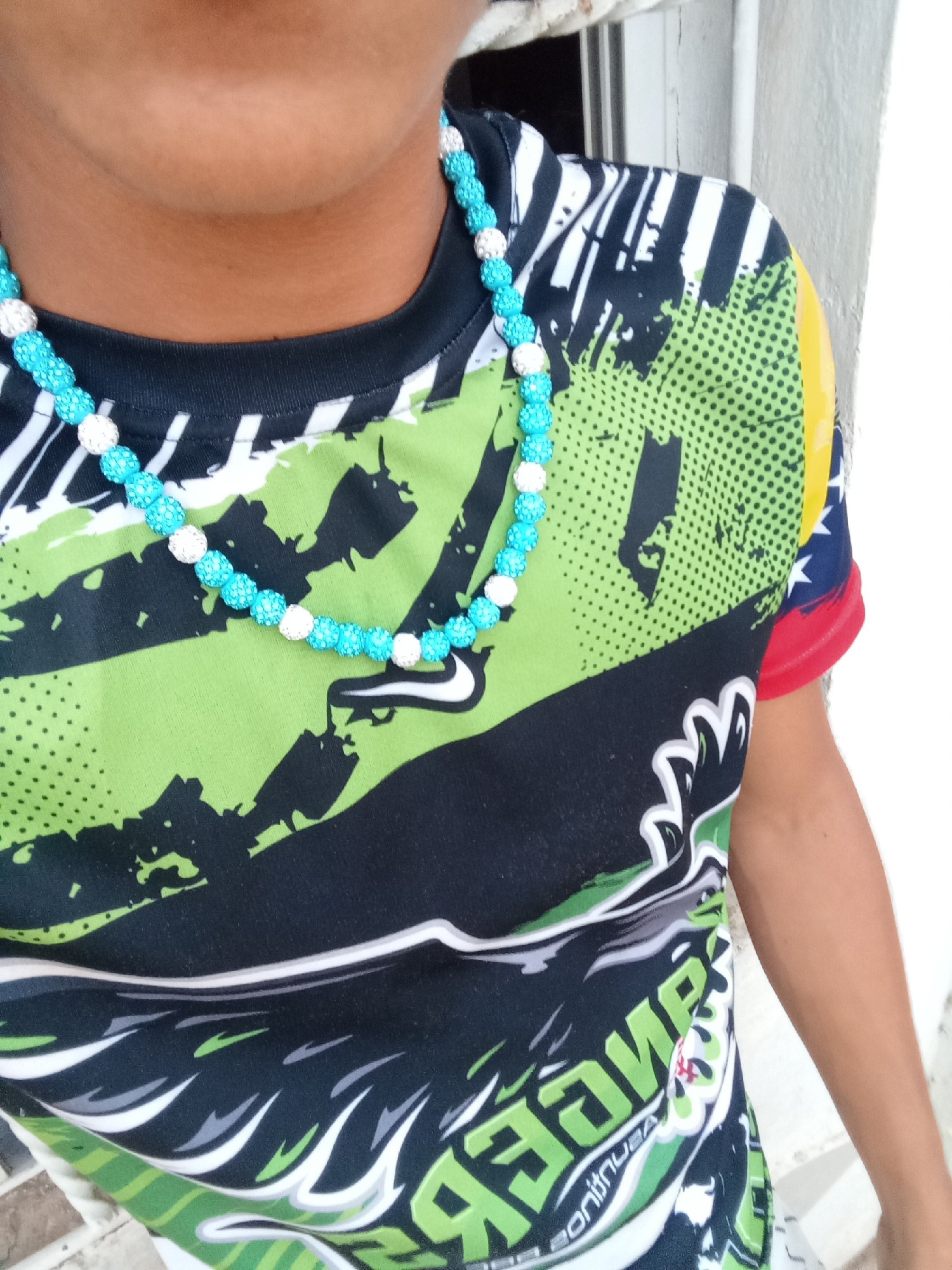 A close-up shot captures the upper torso and neck of a person, likely a child or young individual, set against a plain, light-colored wall. The subject's face is not visible.
The person is wearing a striking beaded necklace composed of alternating light blue and white/silver beads, which appear to have a textured or sparkly surface. The necklace rests over a brightly patterned t-shirt. The t-shirt features a predominantly black base with vibrant green sections, some of which are adorned with a polka-dot pattern. Abstract white and black designs are also integrated into the shirt's graphic.
A notable detail on the right sleeve of the t-shirt is a design resembling the Venezuelan flag, with visible yellow, blue, and red bands, along with small white stars. This detail aligns with the provided location context of Juan Griego, Venezuela. A white Nike swoosh logo is also visible on the shirt, near the necklace. Partially visible, reversed green text on the lower part of the shirt appears to include the words "DANGER" and "CONTINUA."
The lighting is bright and even, suggesting it is daytime. The person appears to be standing, possibly for a photograph, with no specific activity discernible.