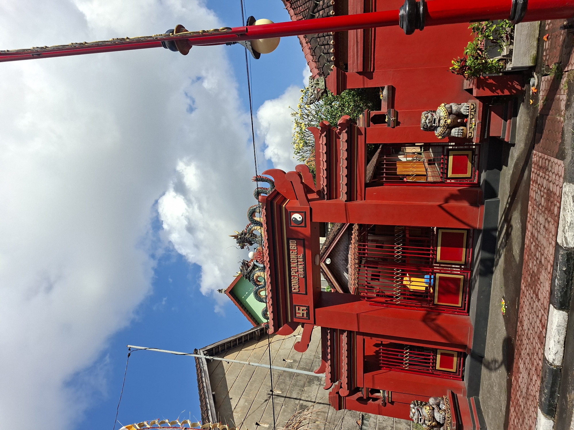 The image shows the bright red entrance to a temple in Gianyar, Indonesia. The temple's gate is ornately decorated with traditional Chinese architectural elements, including curved eaves and dragon statues on the roof. The temple's name, "CONG PO KUNG BIO GIANTAR," is visible above the entrance. Flanking the entrance are two stone lion statues, commonly found in front of temples and palaces. The street in front of the temple is paved with red bricks, and there are potted plants and fallen flowers on the ground. The sky above is a vibrant blue with scattered white clouds, suggesting a sunny day. A red lamppost stands to the right of the temple entrance. The overall scene conveys a sense of vibrant cultural and religious heritage.