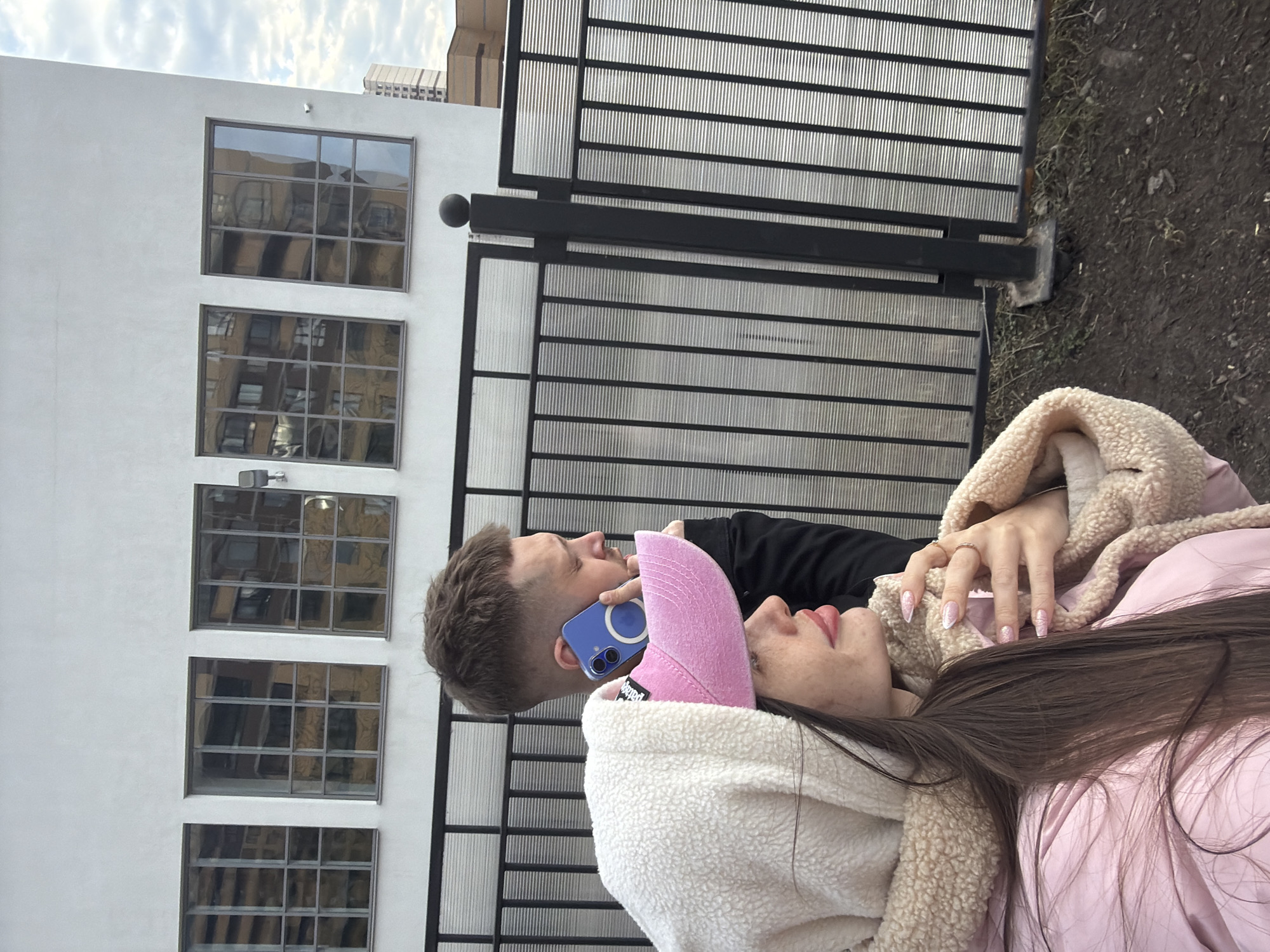 A medium, full shot depicts a man and a woman in front of a building in Saint Petersburg, Russia. The man is standing and holding a blue smartphone to his left ear. His hair is short and brown. The woman is lying on her back, wearing a pink jacket with a tan, plush collar. She is wearing a pink baseball cap. Her hair is long and brown. She is facing to the right, with her head resting near the man's shoulder. Her left hand rests on her chest. The building has several windows, and the sky is cloudy. A black metal gate is behind the couple. The ground is covered in dirt and some grass.