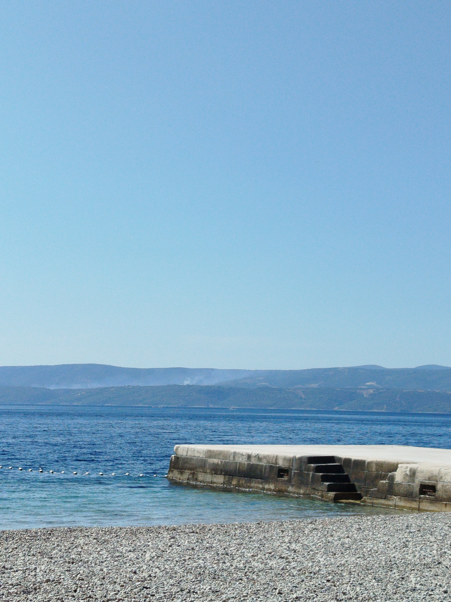 A wide shot captures a serene coastal scene under a clear, pale blue sky. The foreground is dominated by a dense, greyish-white pebble beach. Beyond the beach, the calm, deep blue water of the sea extends towards a hazy, mountainous coastline in the distance. The water near the shore is a lighter, translucent blue, revealing the pebbles beneath. A concrete jetty juts out into the water from the right side of the frame. This jetty features a set of dark steps leading down into the water. A thin rope with evenly spaced white buoys floats on the water's surface, marking a boundary. The distant mountains are softly outlined, with a light atmospheric haze softening their details and the sky above them. There are no people or animals visible in the image.