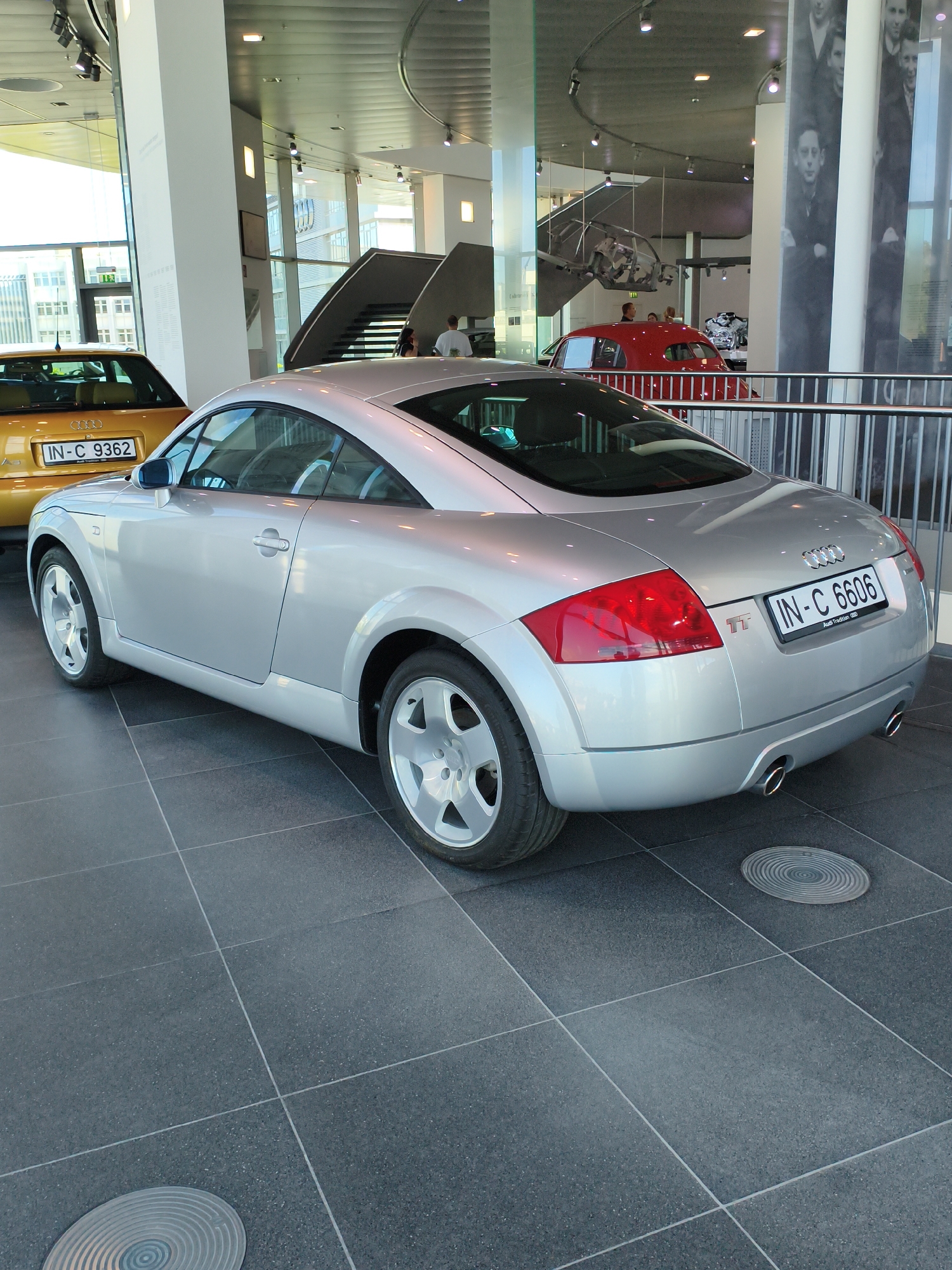 The image shows the interior of a car museum, likely the Audi Museum in Ingolstadt, Germany. The main subject is a silver Audi TT coupe parked on a dark tiled floor. The car is viewed from the rear-right side, showcasing its taillights, exhaust pipes, and license plate "IN-C 6606". To the left of the Audi TT, a yellow car with license plate "IN-C 9362" is partially visible. In the background, a red Audi coupe is on display. The museum features architectural elements like large windows, a staircase, and display panels with images. Several people are casually observing the exhibits, including a man in a white shirt near the red car. The environment suggests a well-lit, modern exhibition space.
