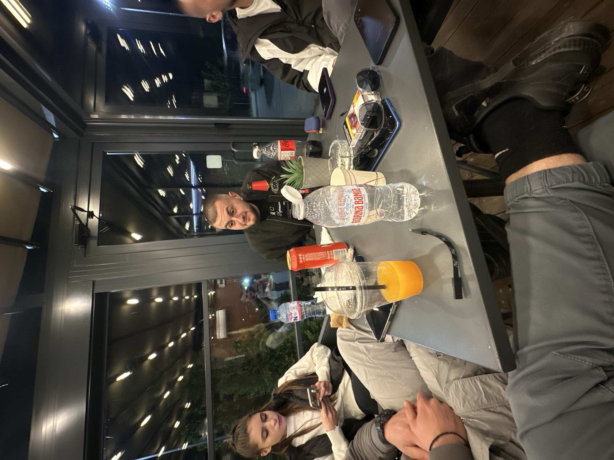 The image depicts a casual nighttime gathering of several people seated around a gray rectangular table, likely in an enclosed outdoor or semi-indoor section of a cafe or restaurant in Plovdiv, Bulgaria. The space is illuminated by overhead strip lighting, which reflects in the large glass panels surrounding the area, indicating it is dark outside.
In the foreground, a man with a short beard and dark clothing looks towards the camera with a neutral expression. To his left, a woman with long brown hair, wearing a white top and a dark vest, is looking down at her smartphone. Partial views of other individuals are visible at the table, including a person wearing a black and white jacket and another whose legs are visible, sporting black Nike socks and black shoes.
The table holds an assortment of drinks and personal items. Visible beverages include a large plastic bottle of "ГОРНА БАНЯ" (Gorna Banya) mineral water, two Coca-Cola bottles (one red, one clear), and a clear plastic cup containing an orange drink, likely orange juice. Other items on the table include multiple smartphones, a pair of sunglasses, a partially visible pack of "Parliament" cigarettes, a small artificial potted plant, and a black charging cable. The overall scene suggests a relaxed social interaction among friends or acquaintances.