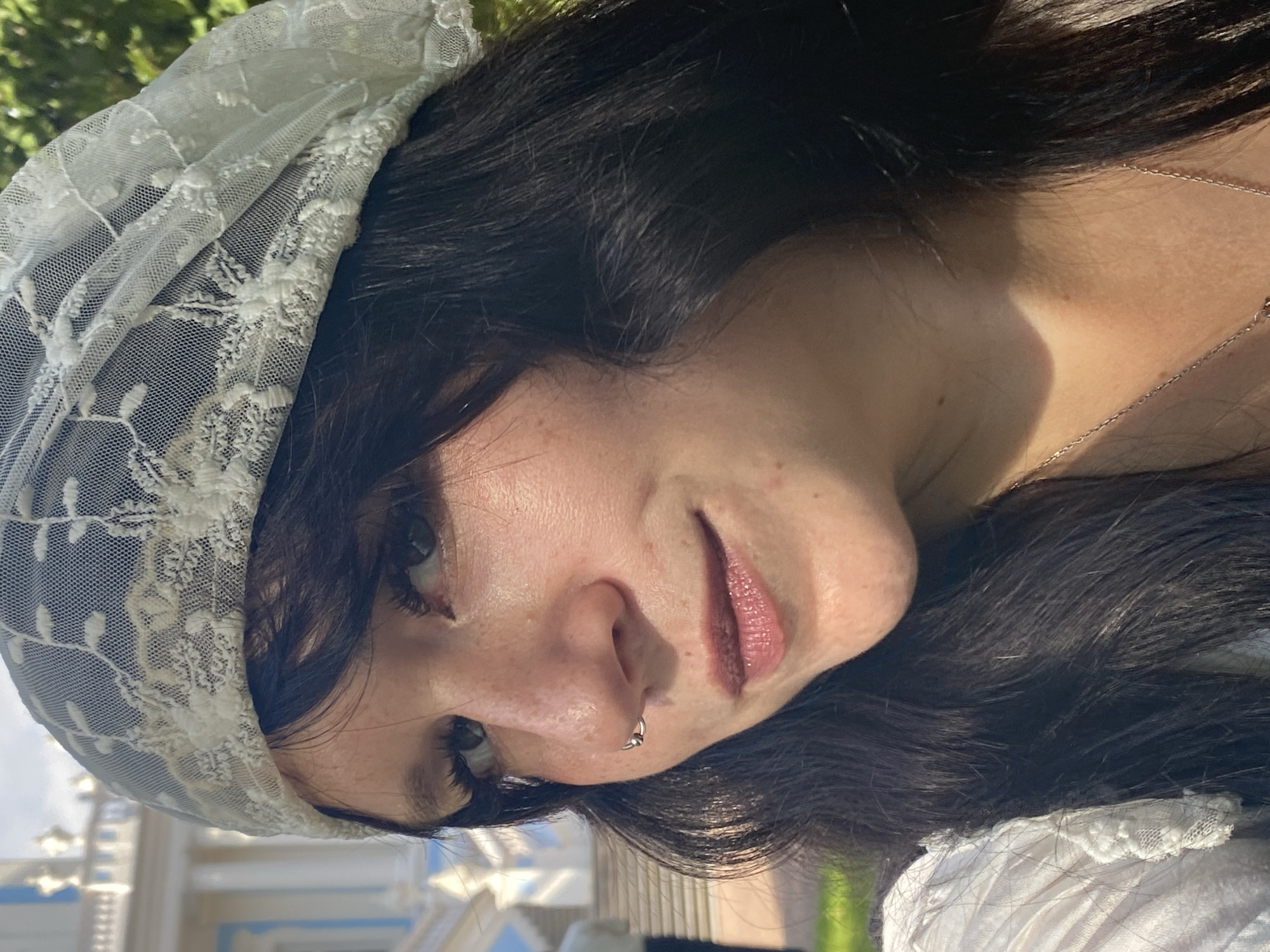 A close-up, outdoor, head-and-shoulders shot shows a young person with dark, wavy hair and fair skin, wearing a white lace headscarf. They are also wearing a white garment with lace trim and a silver necklace. The person has freckles and wears a nose ring. Their eyes are looking to the left of the frame, and their lips are slightly parted. Behind the person, a blurred white building with blue accents and balustrades is visible, consistent with architecture found in the city of Lomonosov, Russia, potentially in the vicinity of the Catherine Palace or its park. Green foliage is also visible in the background, suggesting a sunny day. The lighting indicates natural daylight.