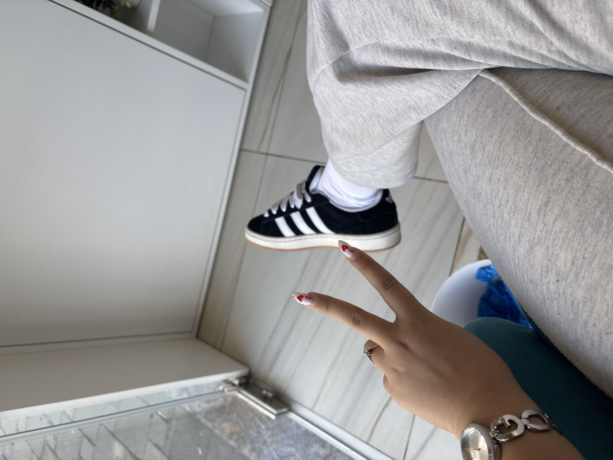 A person wearing gray sweatpants and a black Adidas sneaker with white stripes is sitting indoors on what appears to be a dark green surface. The person's hand is raised in a peace sign, revealing a watch and painted fingernails with heart designs. A white cabinet is visible to the left. The location is unknown and there is no indication that this is in Plovdiv, Bulgaria.