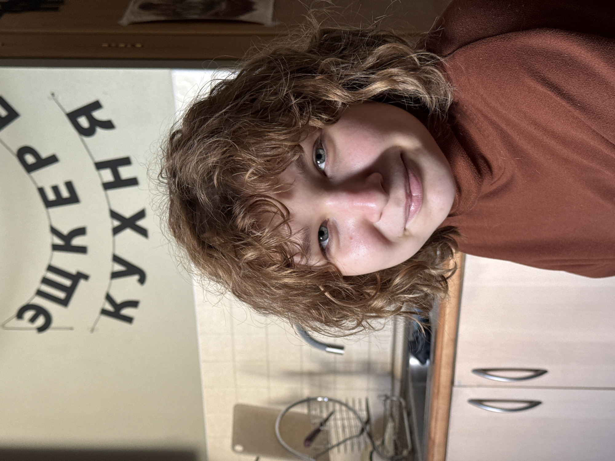 This is a close-up, overhead shot of a young person with curly, light brown hair, wearing a rust-colored turtleneck. Their face is turned towards the camera, with green eyes, a slight smile, and freckles across their nose and cheeks. The lighting suggests an indoor setting, possibly a kitchen, given the visible cabinets and a countertop with some kitchen utensils.
To the left of the person, on a white surface, is text in black, Cyrillic lettering arranged in a circular pattern. The visible words appear to be "ЕЩЁ КУХНЯ," which translates to "MORE KITCHEN." The text is oriented vertically.
The overall mood is casual and relaxed, with the subject looking directly at the camera. The image seems to have been taken in a domestic environment, likely within Saint Petersburg, Russia, based on the provided location context. There are no indications of the time of day or weather conditions outside.