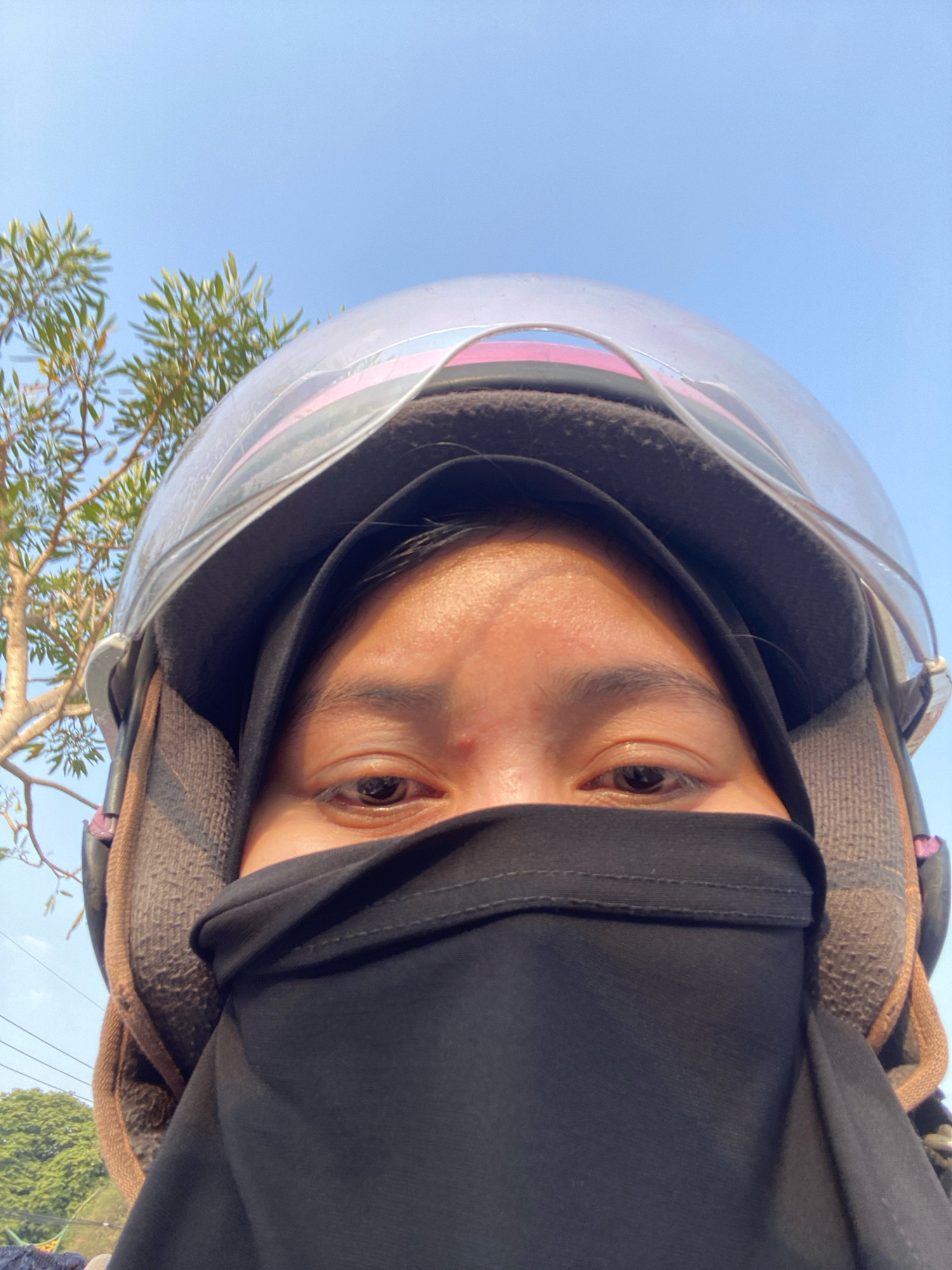 A close-up, low-angle selfie captures a person wearing a black hijab and a motorcycle helmet against a clear blue sky. The person's eyes are visible above the black fabric covering their nose and mouth. A faint pink stripe is visible on the helmet's interior lining. To the left of the helmet, a tree with sparse, green foliage is partially in frame. In the upper left corner, the sky is a pale blue, transitioning to a slightly darker blue towards the top of the image. The background suggests an outdoor, possibly urban, setting with some power lines visible near the bottom right. The focus is on the subject's face and the protective gear.