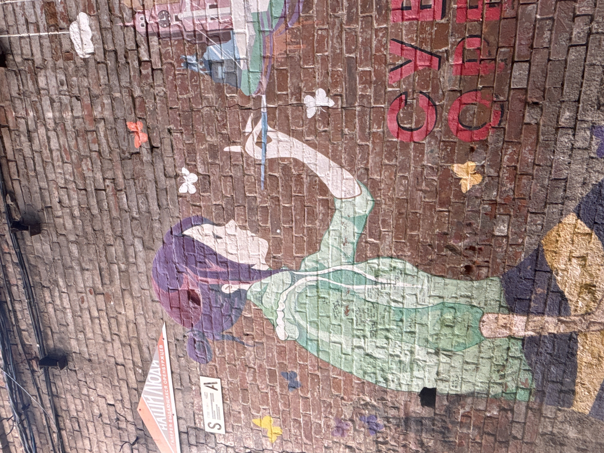 The image shows a mural on a brick wall in Orenburg, Russia. The mural features a person with purple hair and a green dress. Their right arm is raised, reaching toward a blue object. There are butterfly shapes of various colors surrounding the figure. Red letters are visible above the person. A sign with Russian text is partially visible on the lower left corner of the image. The environment appears to be outdoors, with wires visible in the upper left.