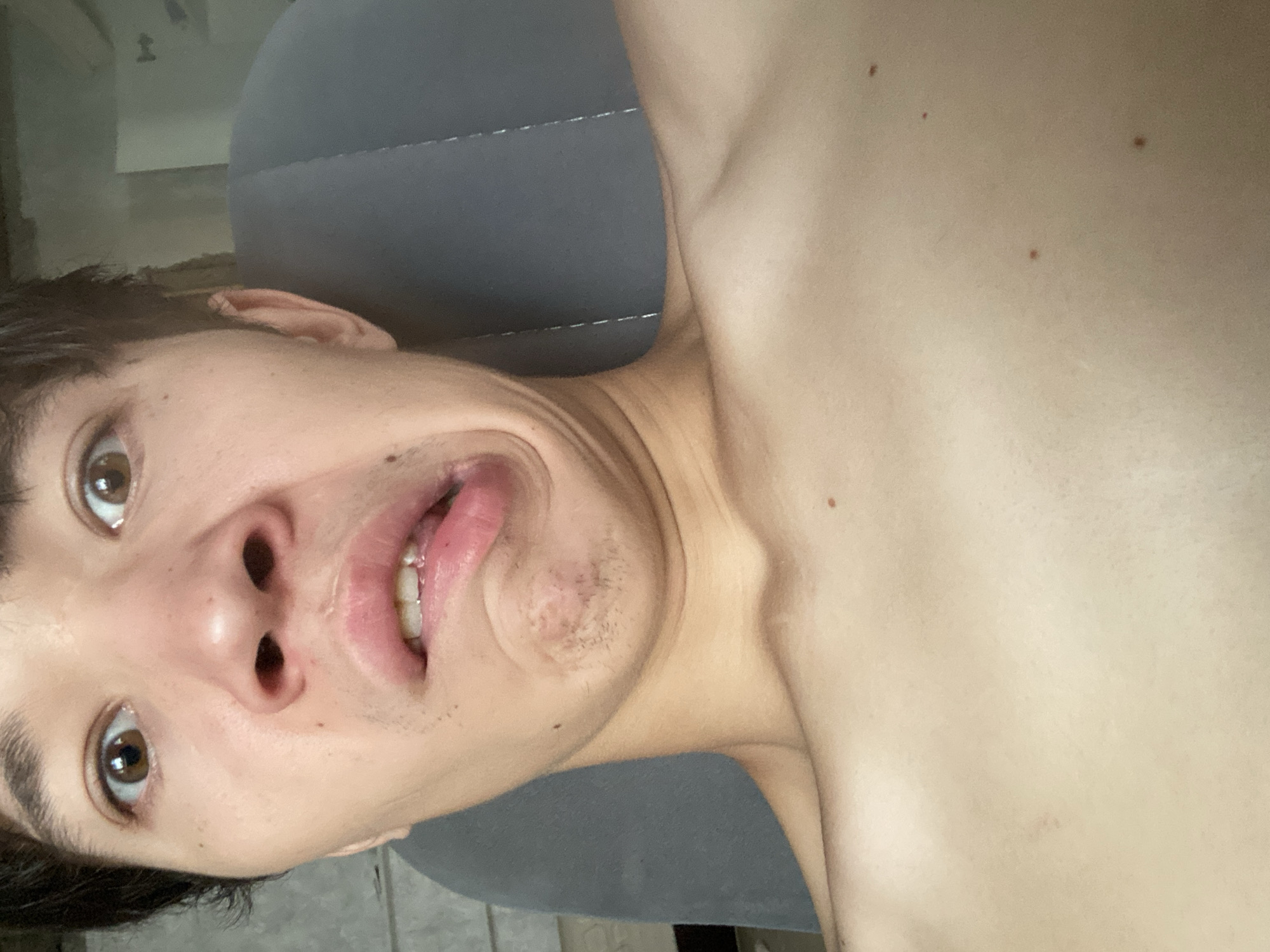The image is a close-up, selfie-style photograph of a young person's face and upper chest. The person has brown eyes and dark hair. Their facial expression is distorted, with one side of their mouth pulled up and the other down, revealing their teeth. There is some light stubble on their chin. The person is shirtless, and their skin is visible. In the background, a gray upholstered surface, likely a car seat or chair, is visible. The lighting is somewhat harsh, casting shadows on the person's face and chest. The image does not contain any elements that would suggest a location in Shakhtinsk, Kazakhstan.