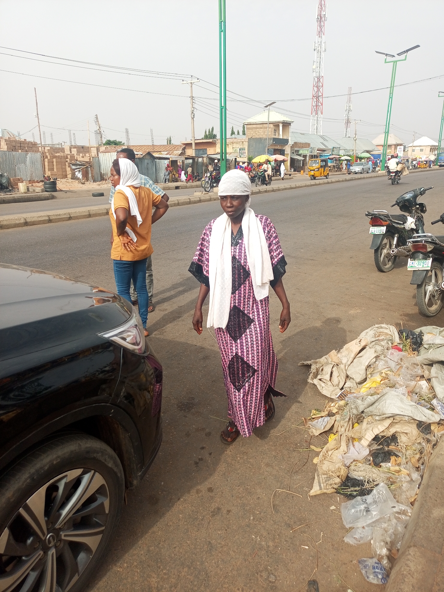 The image depicts a scene in what appears to be a bustling area of Tempure, Nigeria. A woman wearing a patterned, purple and pink dress and a white headscarf walks on the shoulder of a paved road. A white cloth drapes over her shoulders. To her right is a large pile of garbage consisting of plastic bags, organic waste, and various debris.
A black car occupies the left side of the frame, partially obscuring another person standing on the side of the road with another person. Two motorcycles are parked near the pile of garbage, their license plates partially visible.
In the background, there are buildings, people walking along the sidewalk, motorcycles, and auto rickshaws ("Keke Napep"). Utility poles and communication towers are visible in the distance against a somewhat hazy sky. The overall environment suggests a busy, urban setting with sanitation challenges.