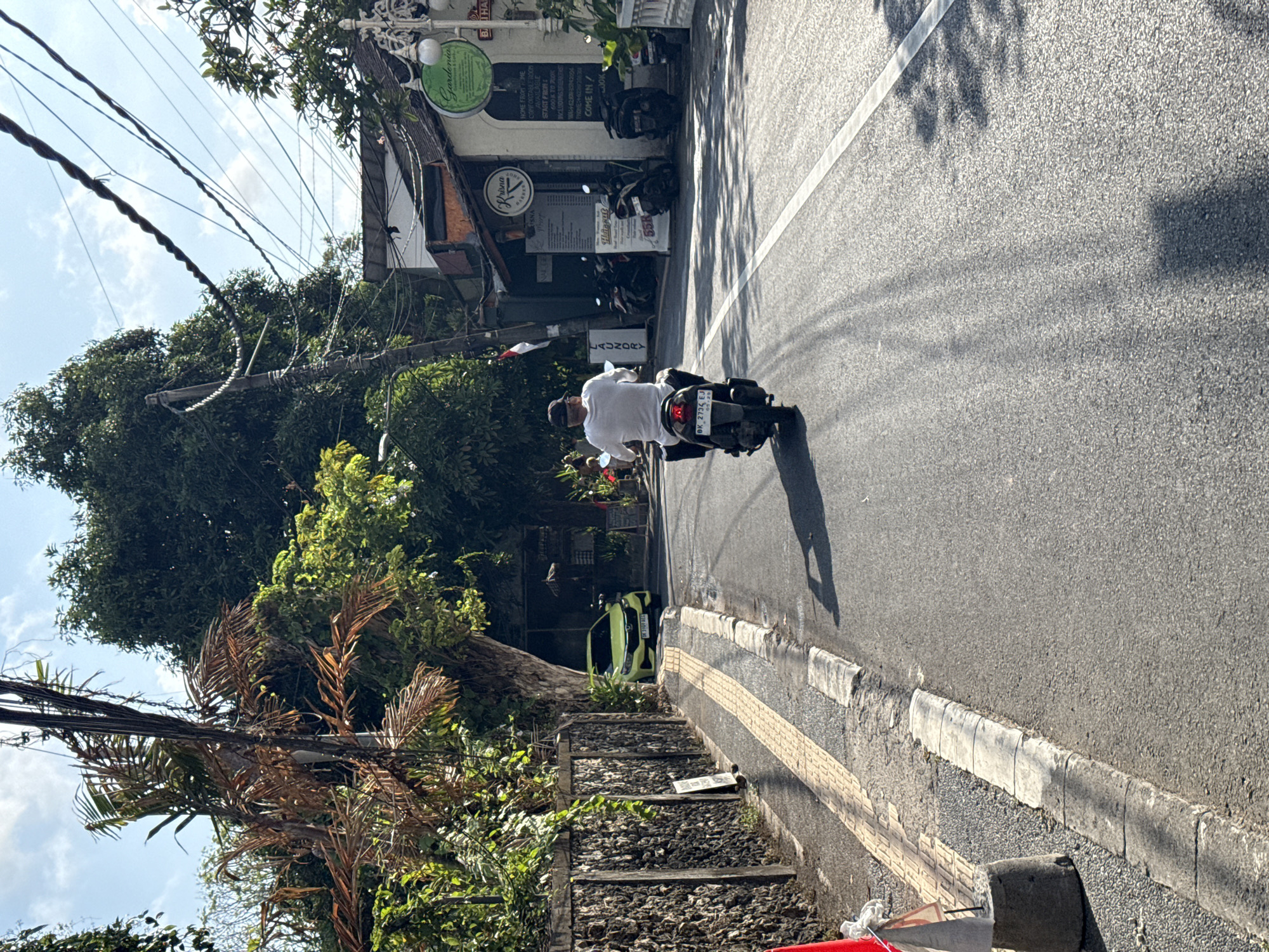 An eye-level, medium shot captures a man riding a scooter down a paved road in Denpasar, Indonesia. The scooter, dark gray in color, is moving away from the camera. The rider, wearing a white t-shirt, black pants, and a dark cap, is partially obscured by the scooter. The road is asphalt with a white line marking the lane. To the left, a stone wall lines the road, followed by a sidewalk with yellow tactile paving. Beyond the wall, lush green foliage and trees are visible. In the background, several shops with signs are present, one with a green circular logo that reads "Gardenia". Power lines crisscross overhead, and large trees provide shade in some areas. A bright green car is parked on the side of the road near the shops. The sky is blue with scattered white clouds. The scene is illuminated by bright sunlight, casting shadows on the road.