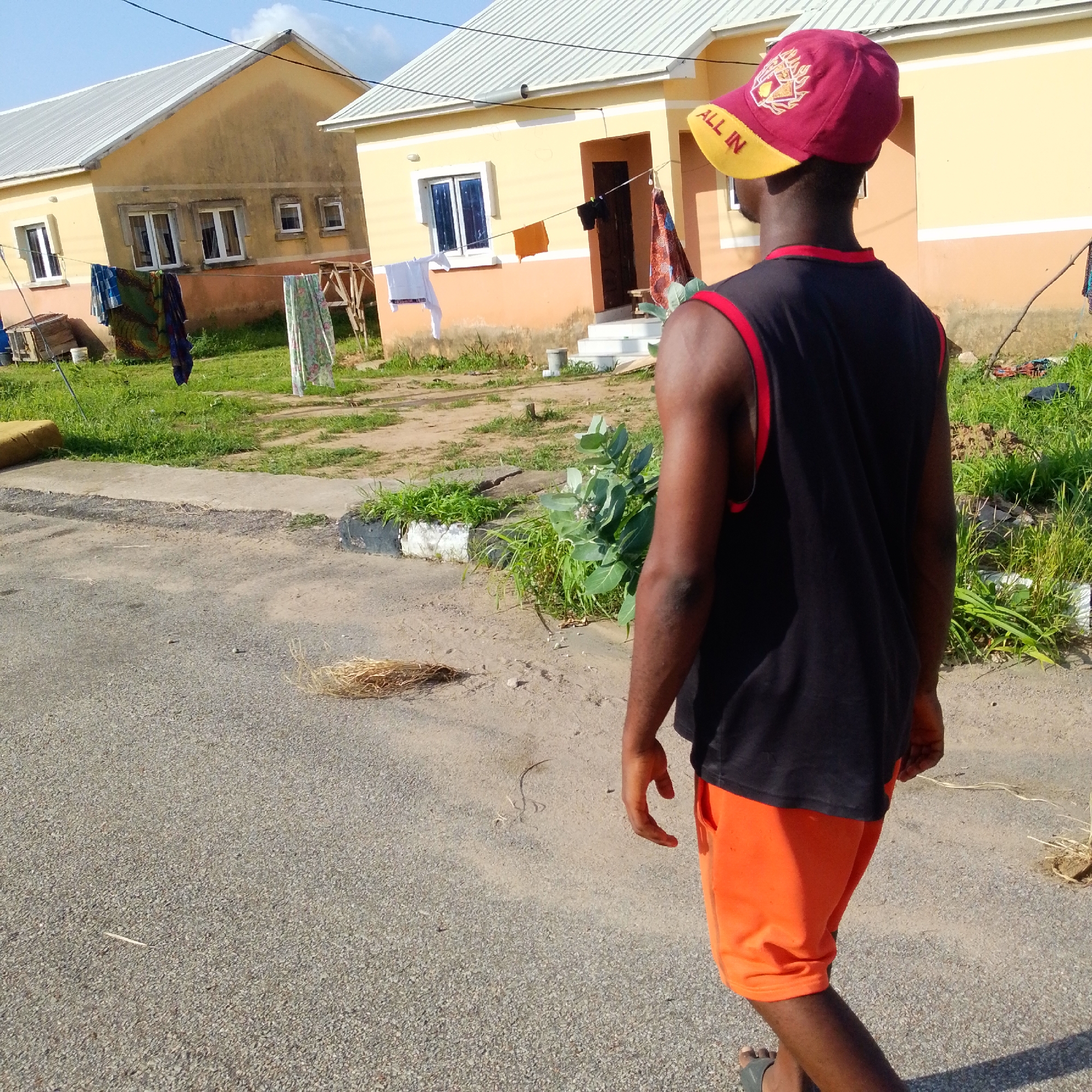 In an outdoor scene in an unknown city, a person walks on a paved road. The person is seen from behind, wearing a black tank top with red trim and orange shorts. They are also wearing a red and yellow baseball cap. The background consists of two residential buildings, one yellow and one with a tan lower half and white upper half, with laundry hanging on lines in front of them. Green grass and vegetation are present around the buildings and along the side of the road. The ground is a mix of asphalt and dirt. The sky is visible, with some clouds.