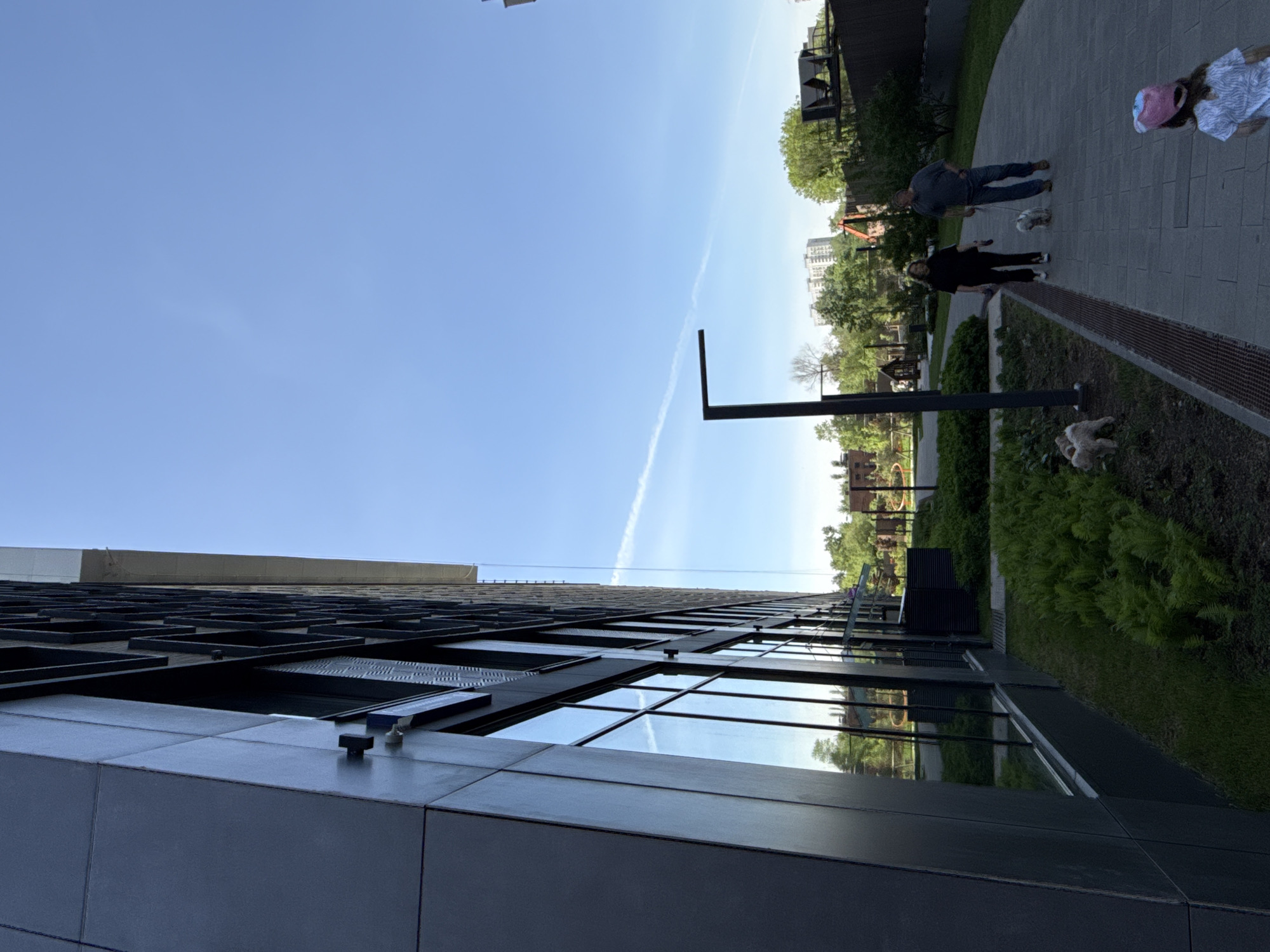 The image shows a view from the base of a modern building with a grid-like facade. The sky is blue with a contrail visible. Adjacent to the building, there's a landscaped area with a paved path. Two adults walk on the path accompanied by small dogs. Additional dogs appear to be resting in the planted area next to the path. Buildings and trees are visible in the background. The image appears to have been taken near the High Park residential complex, Moscow, Russia.