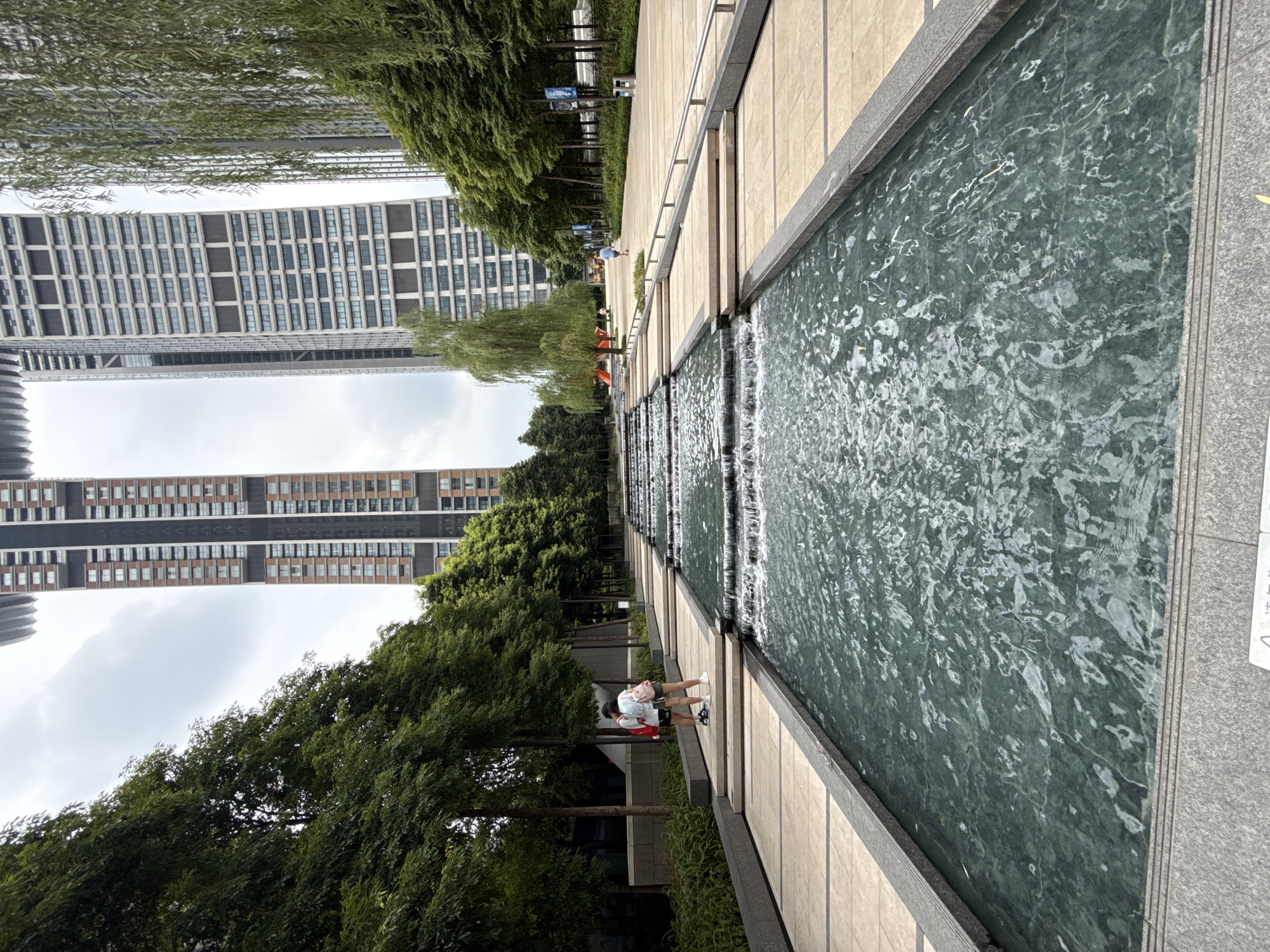 A wide-angle, eye-level shot captures a modern urban park scene in Yuzhong District, China. A multi-tiered water feature with cascading waterfalls dominates the foreground, its dark green water reflecting the overcast sky. The water feature is bordered by a paved walkway with light-colored tiles and dark stone edges.
To the left of the water feature, two individuals, a woman with short dark hair wearing a red top and denim shorts, and another person in a light-colored top and shorts, stand near a tree-lined path. They are facing away from the camera and appear to be interacting. Further into the scene, a man in a blue shirt and shorts walks along the paved path, and another person is visible in the distance near a red slide.
Lining the path are dense green trees and tall modern buildings. On the left, a tall, reddish-brown skyscraper with numerous windows stands prominently. On the right, a lighter-colored, more utilitarian skyscraper rises, its facade also covered in windows. In the background, more trees and buildings complete the urban landscape. The sky is heavily overcast, suggesting a cloudy day. The overall impression is one of a well-maintained, contemporary urban public space.